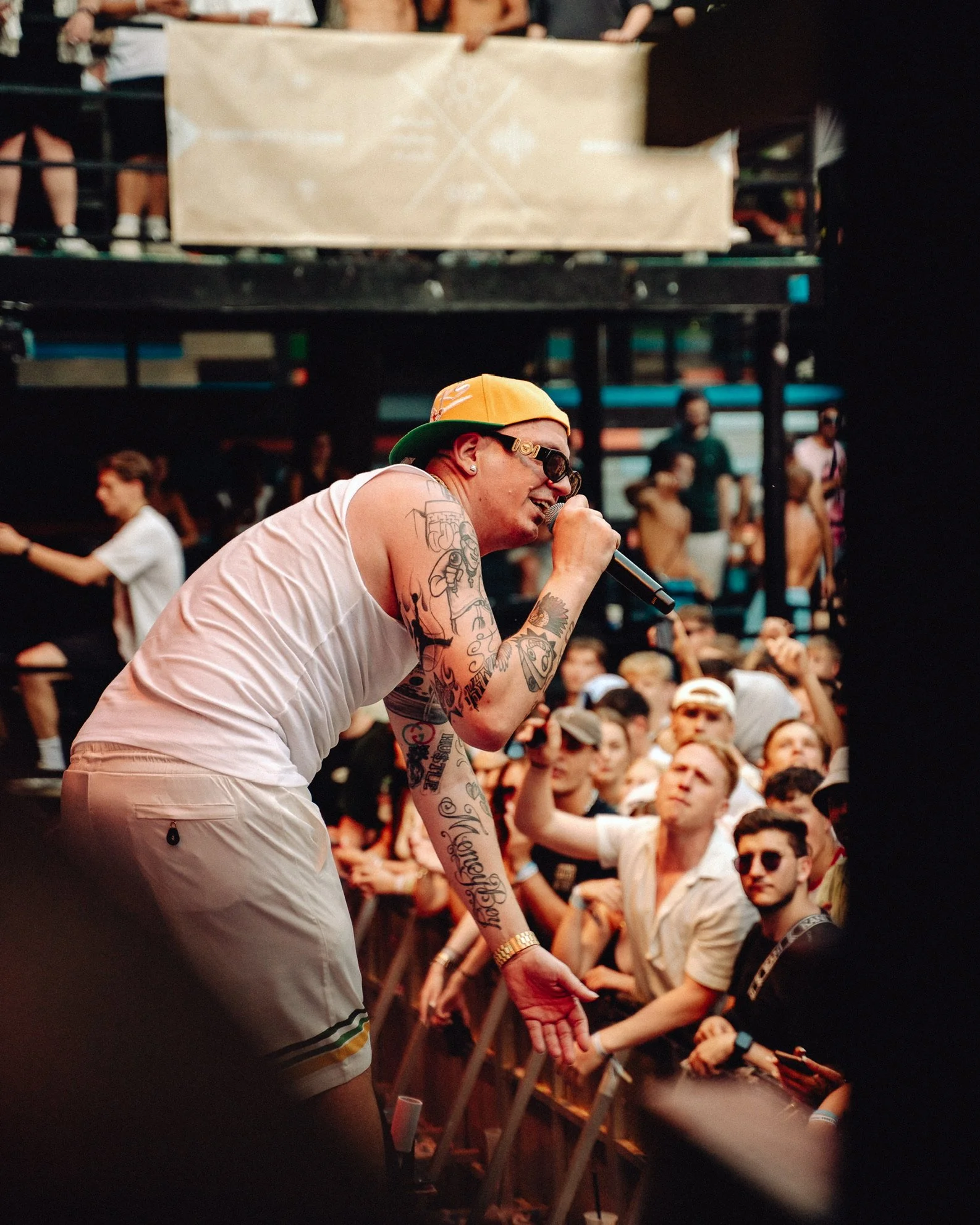 Ein junger Sänger mit Tattoos, eine gelbe Cap, Sonnenbrille und weißem Tanktop singt auf der Bühne vor einem begeisterten Publikum bei einem Konzert.