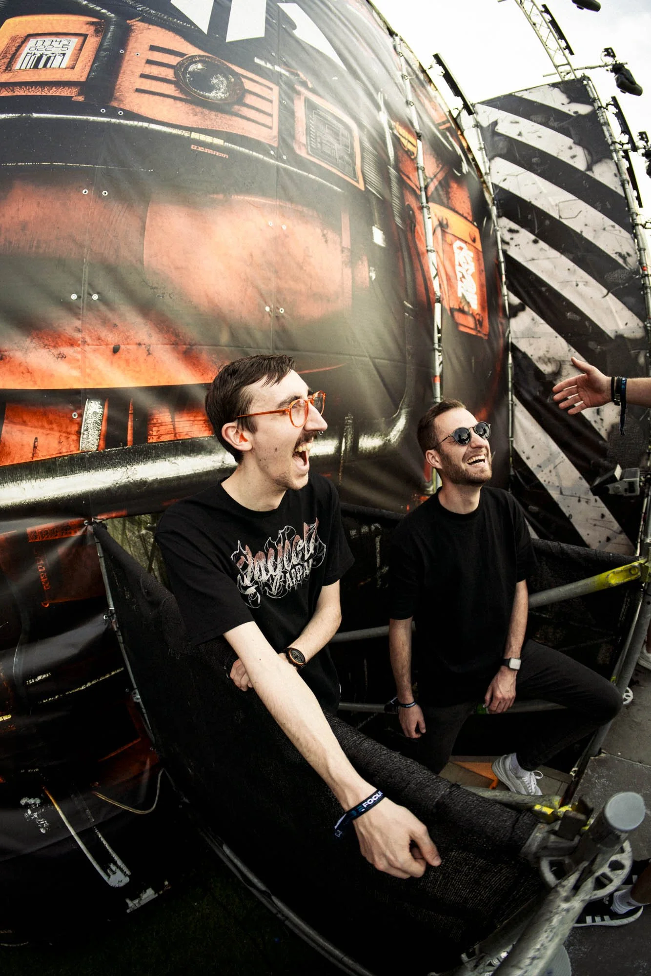 Zwei lachende Männer sitzen auf einem Throne bei einem Festival, im Hintergrund ein großes Bühnenbild mit einem Feuerwehrauto-Design.