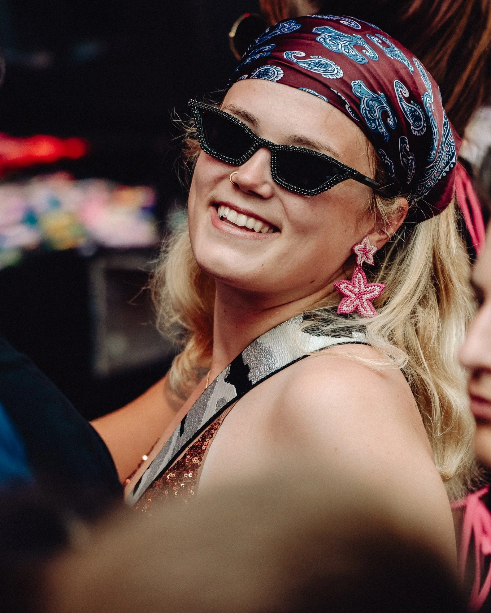 Junge Frau trägt Sonnenbrille, Bandana, große rosa Starfish-Ohrringe und ein glitzerndes Outfit, lächelt bei einer Feier.