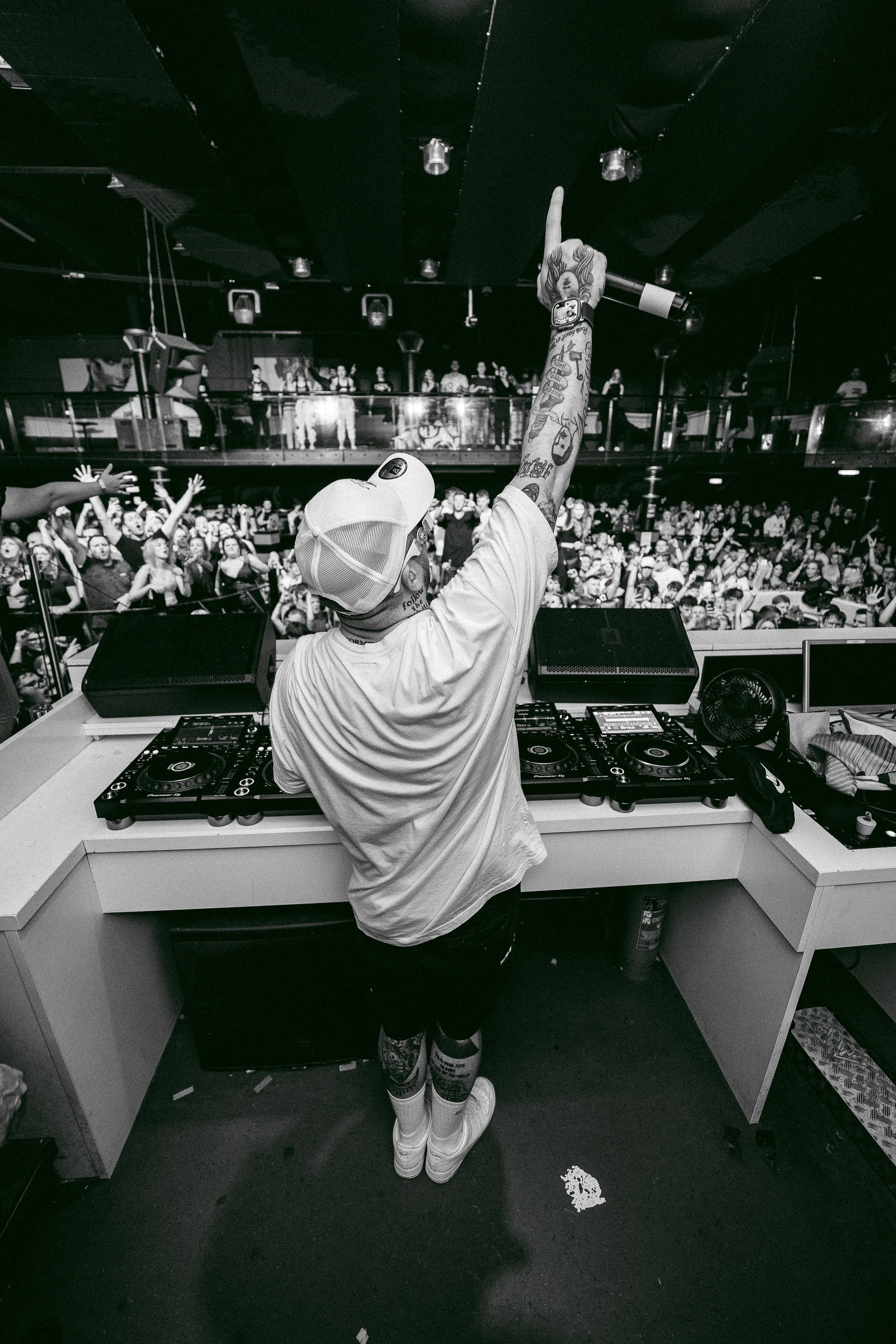 Ein DJ in einer Diskothek, der vor einem Publikum auflegt, mit ausgestrecktem Arm nach oben und einem weißen Cap.