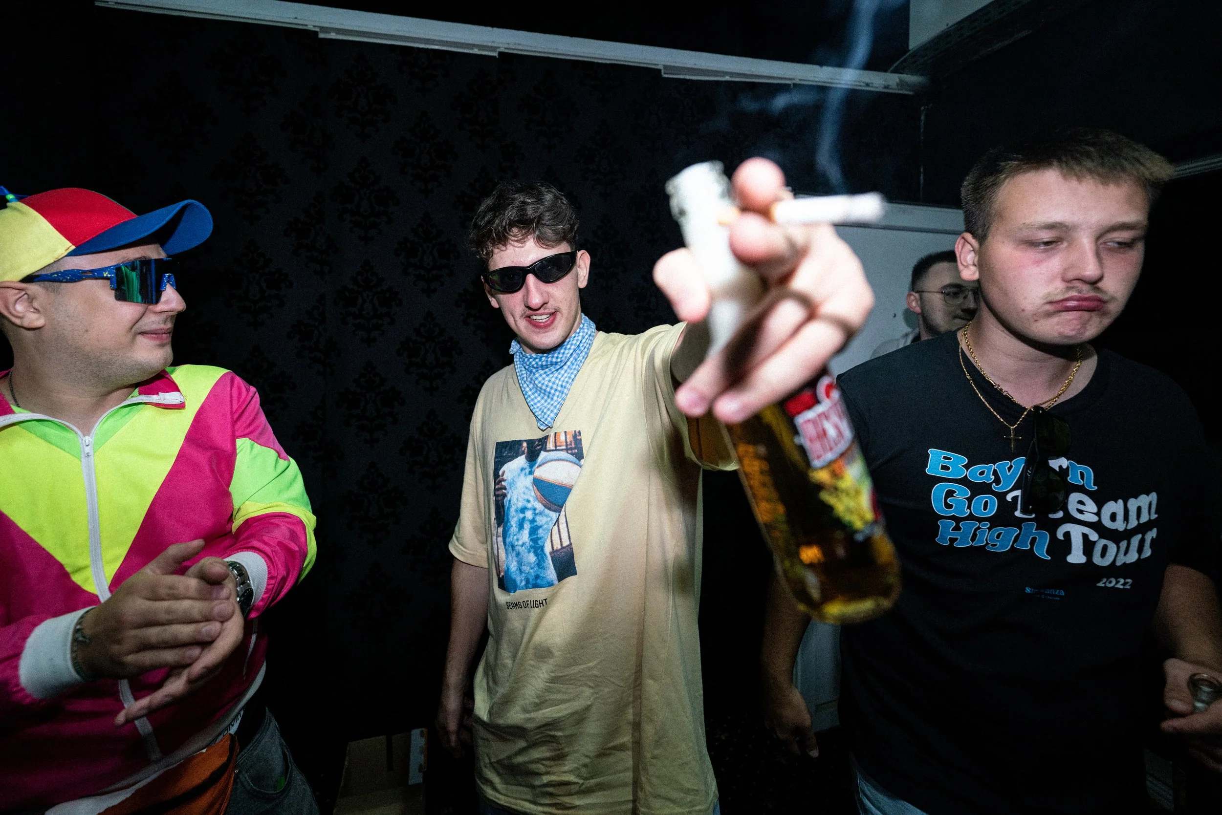 Junge Männer in einem Club, einer hält eine Zigarette, einer trägt eine Sonnenbrille, alle wirken entspannt.