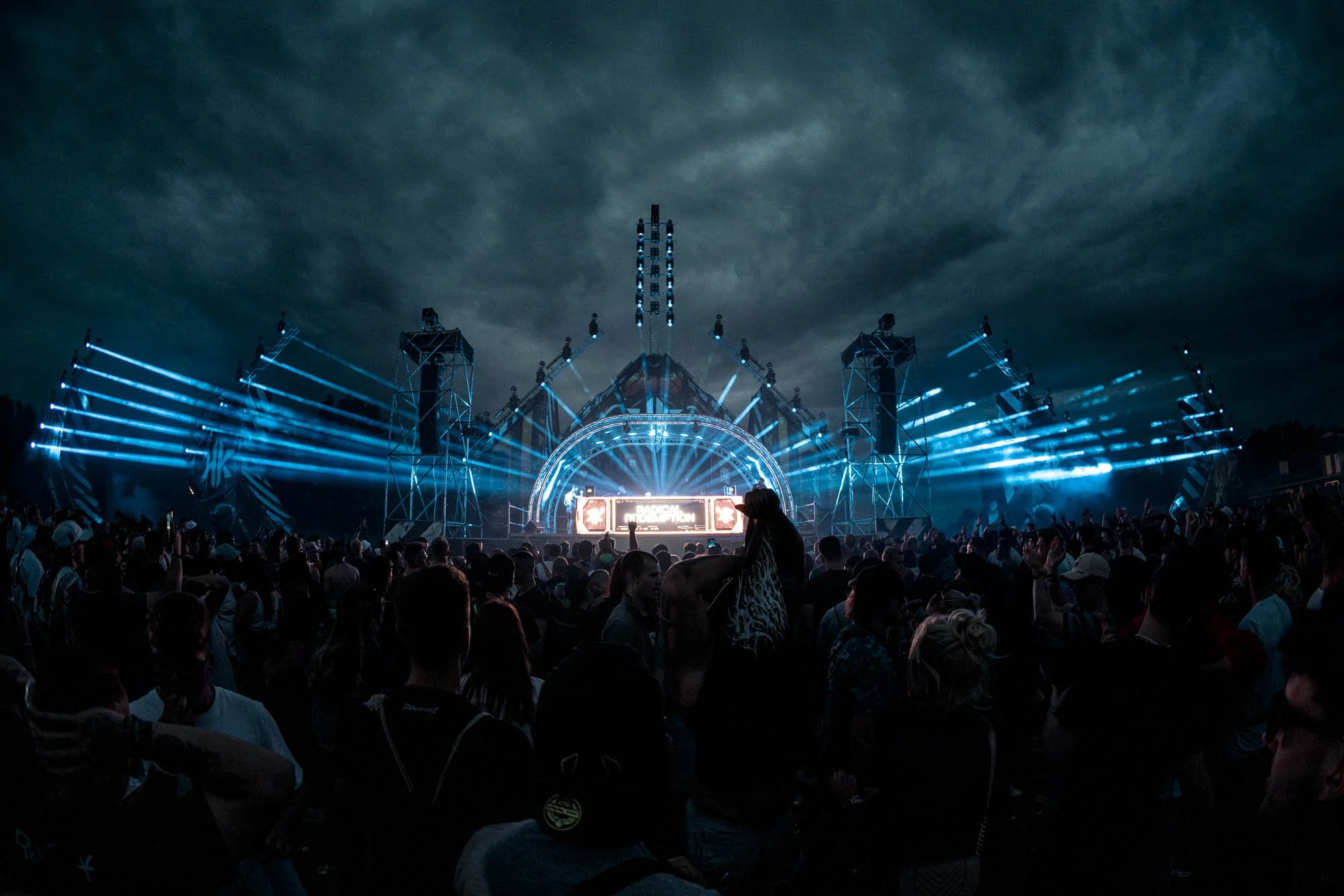 Eine große Menschenmenge auf einem Open-Air-Musikfestival bei Nacht, mit einer Bühne und blauen Lichtinstallationen gegen einen dunklen Himmel.