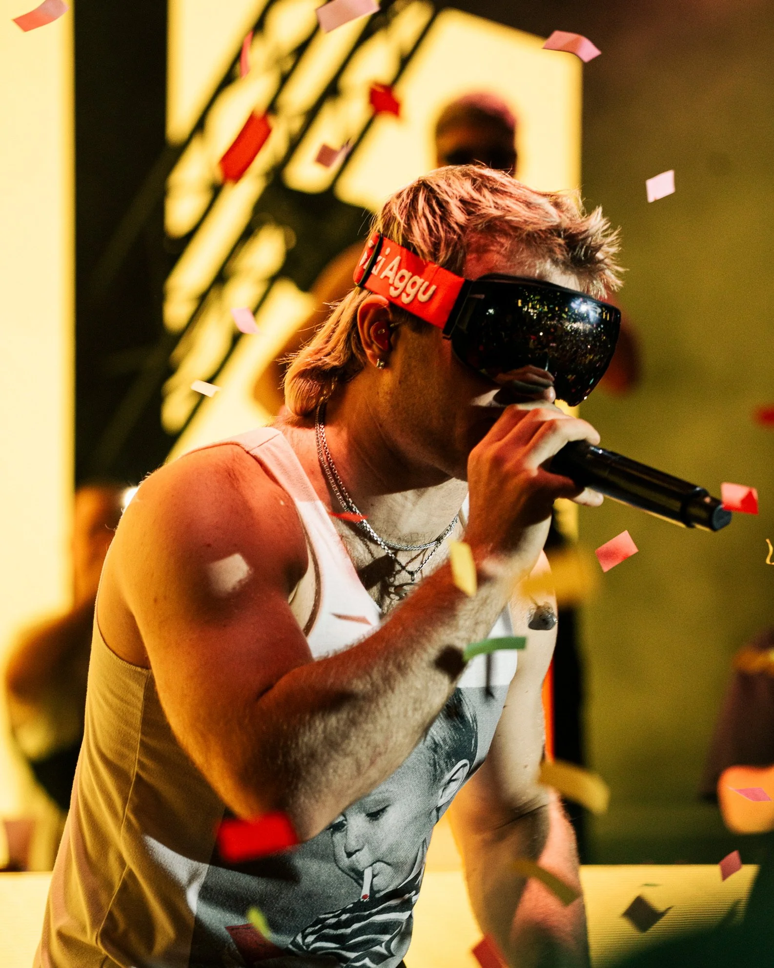 Ein junger männlicher Sänger mit rebellischer Frisur trägt eine LED-Brille und ein Tanktop mit einem Baby-Foto, während er auf einem Konzert singt, umgeben von Konfetti.