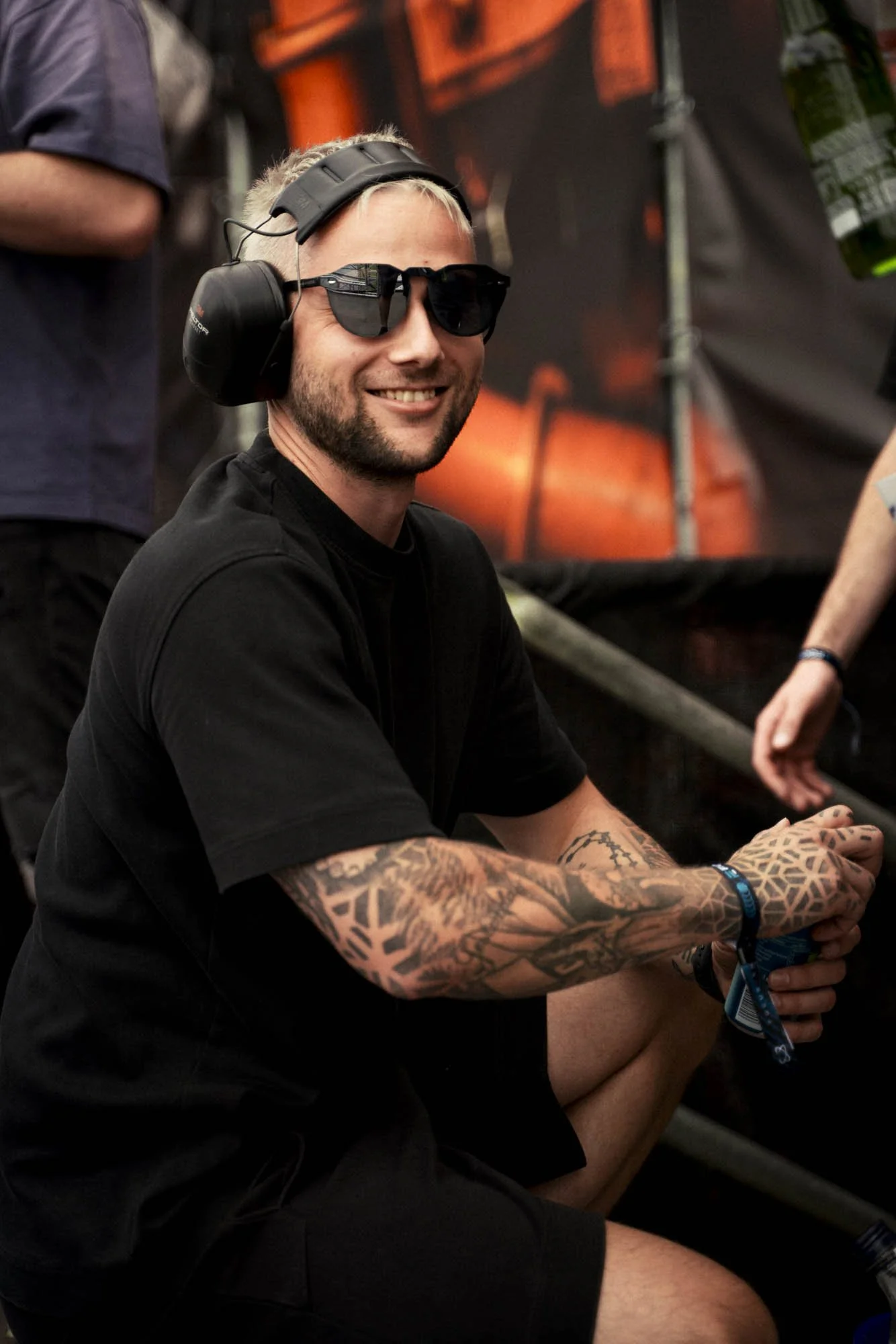 Junge Person mit schwarzen T-Shirt, Sonnenbrille, Kopfhörer mit Mikrofon, Tattoo-Arm und Lanyard, lächelnd auf einem Festival oder Konzert.