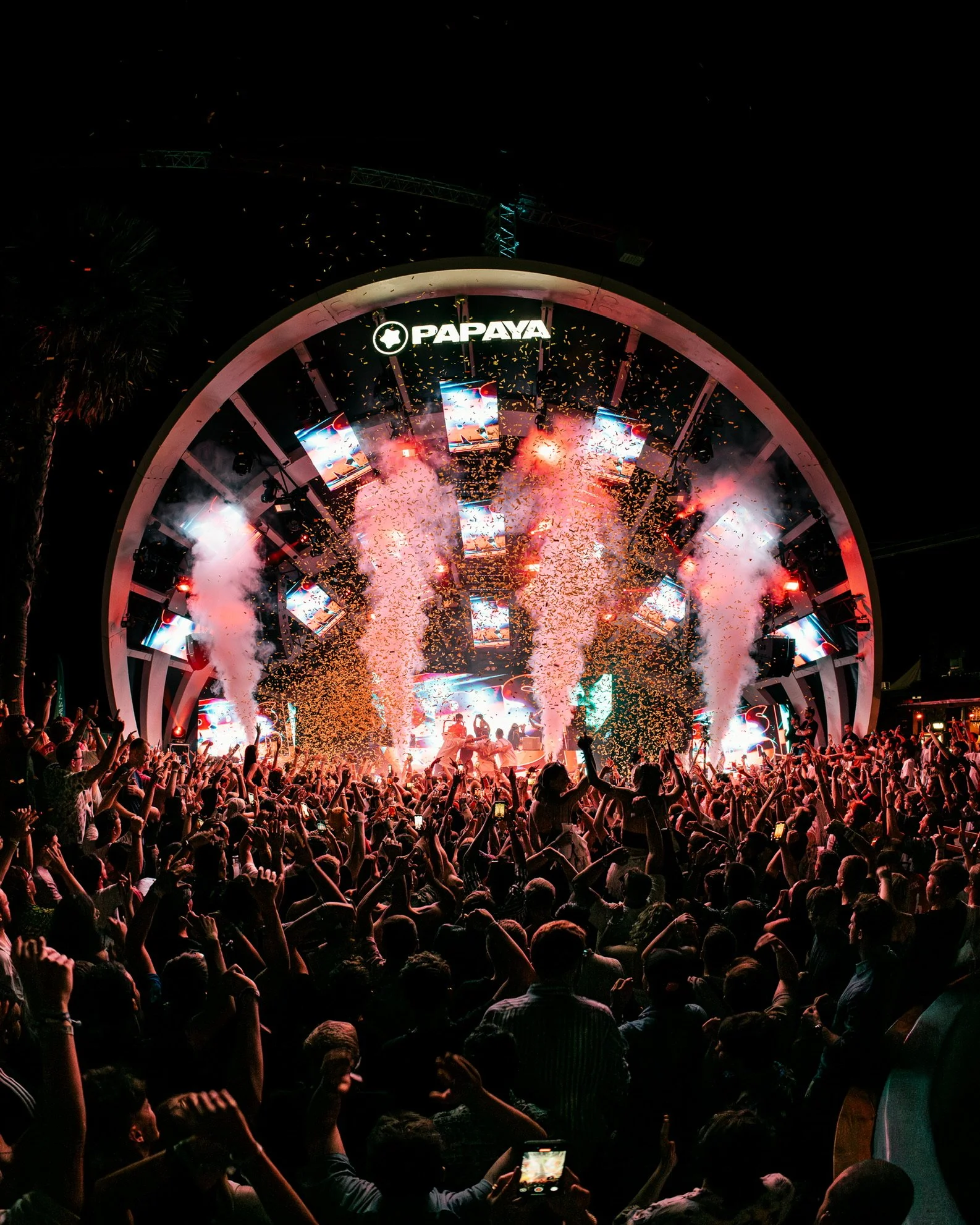 Menschen feiern bei einem Konzert auf eine große Bühne mit Lichtern, Rauch und Konfettiregen, das Logo 'PAPAYA' ist sichtbar.