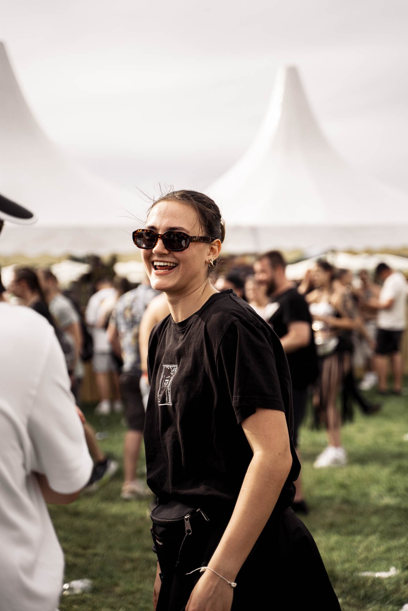 Eine lachende junge Frau mit dunkler Sonnenbrille, dunklem T-Shirt und schwarzer Tasche, die auf einem Festival im Freien unter einem großen weißen Zelt ist, umgeben von Menschen.