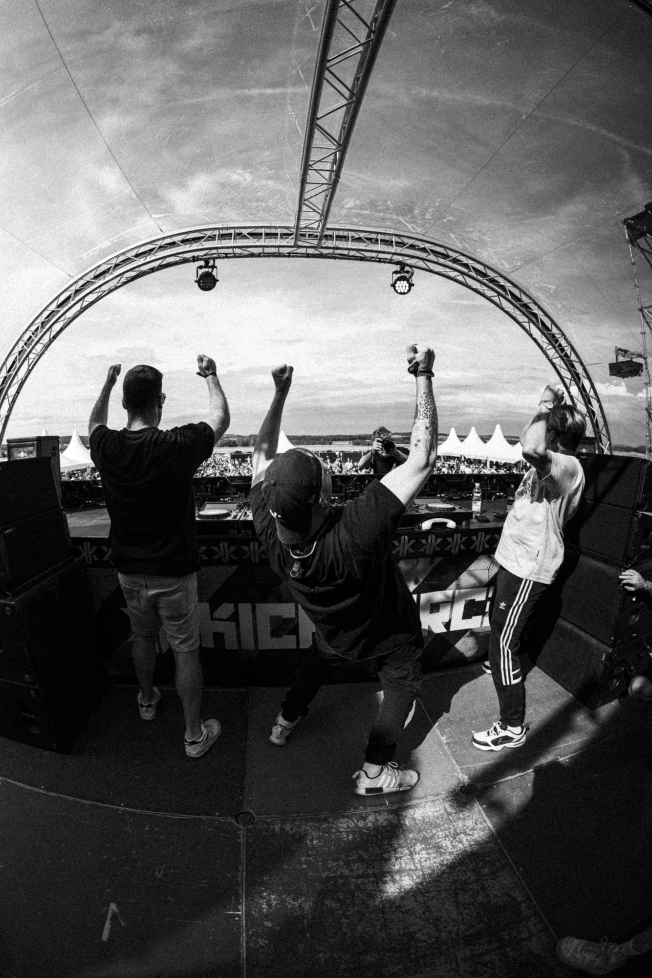 Drei Personen auf einer Bühne, die zum Publikum tanzen und feiern bei einem Open-Air-Festival, im Hintergrund ein beschirmter Himmel und Zelte.