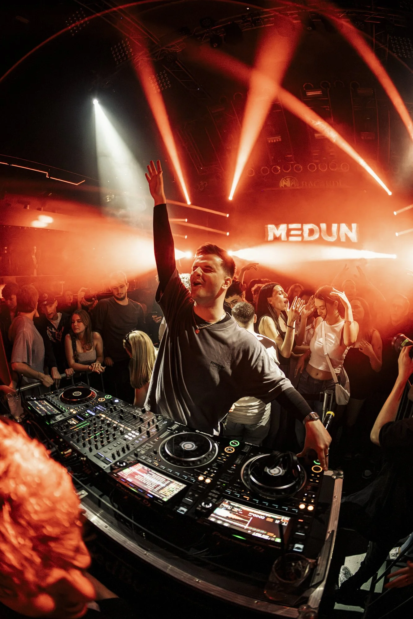 DJ beim Auflegen auf einer Party mit tanzenden Menschen, rote Licht- und Lasereffekte, im Hintergrund ein Bildschirm mit dem Schriftzug 'MEDUN'.