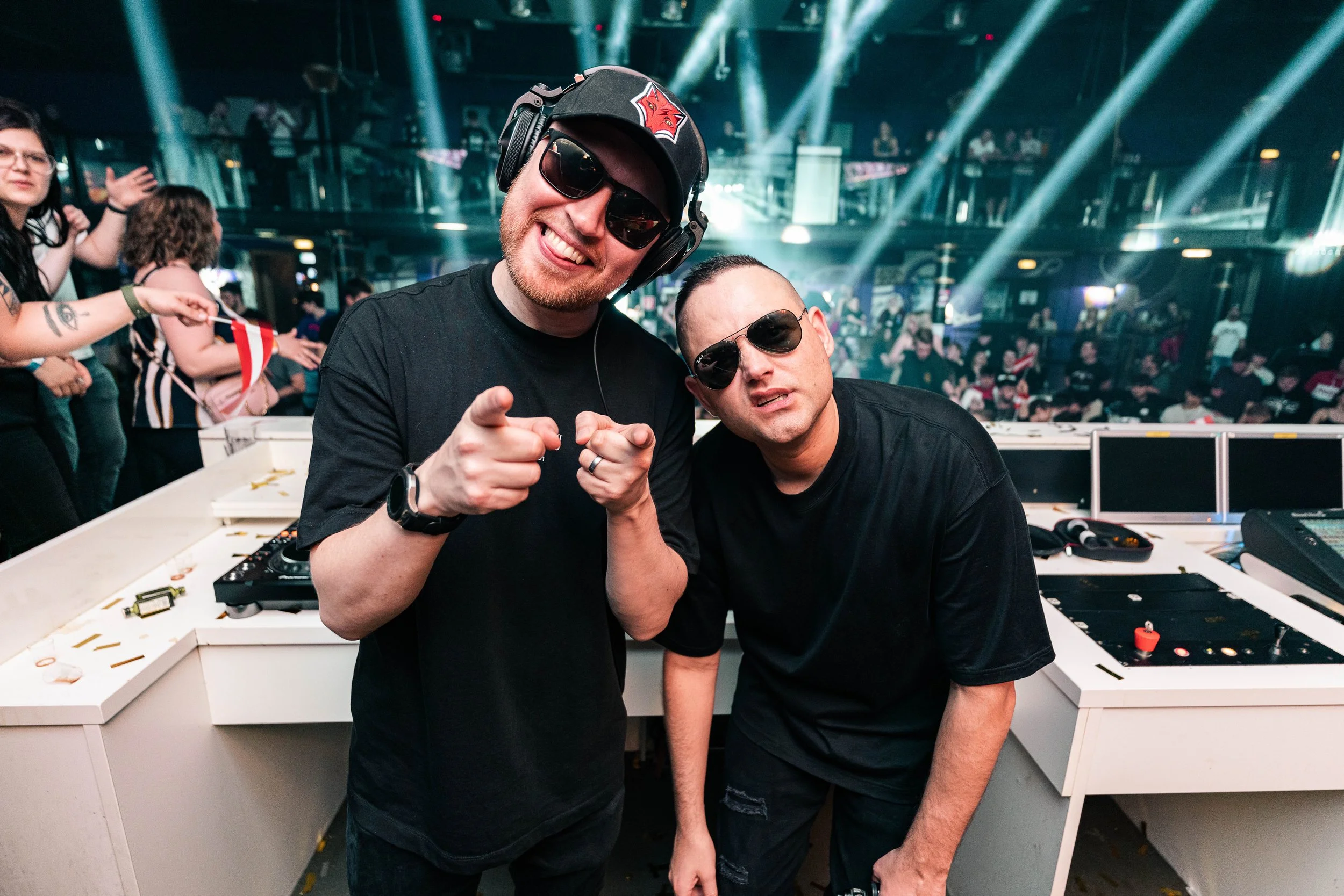 Zwei Männer mit Sonnenbrillen hinter einem DJ-Pult in einem Club, eine Party-Szene mit tanzenden Menschen im Hintergrund.