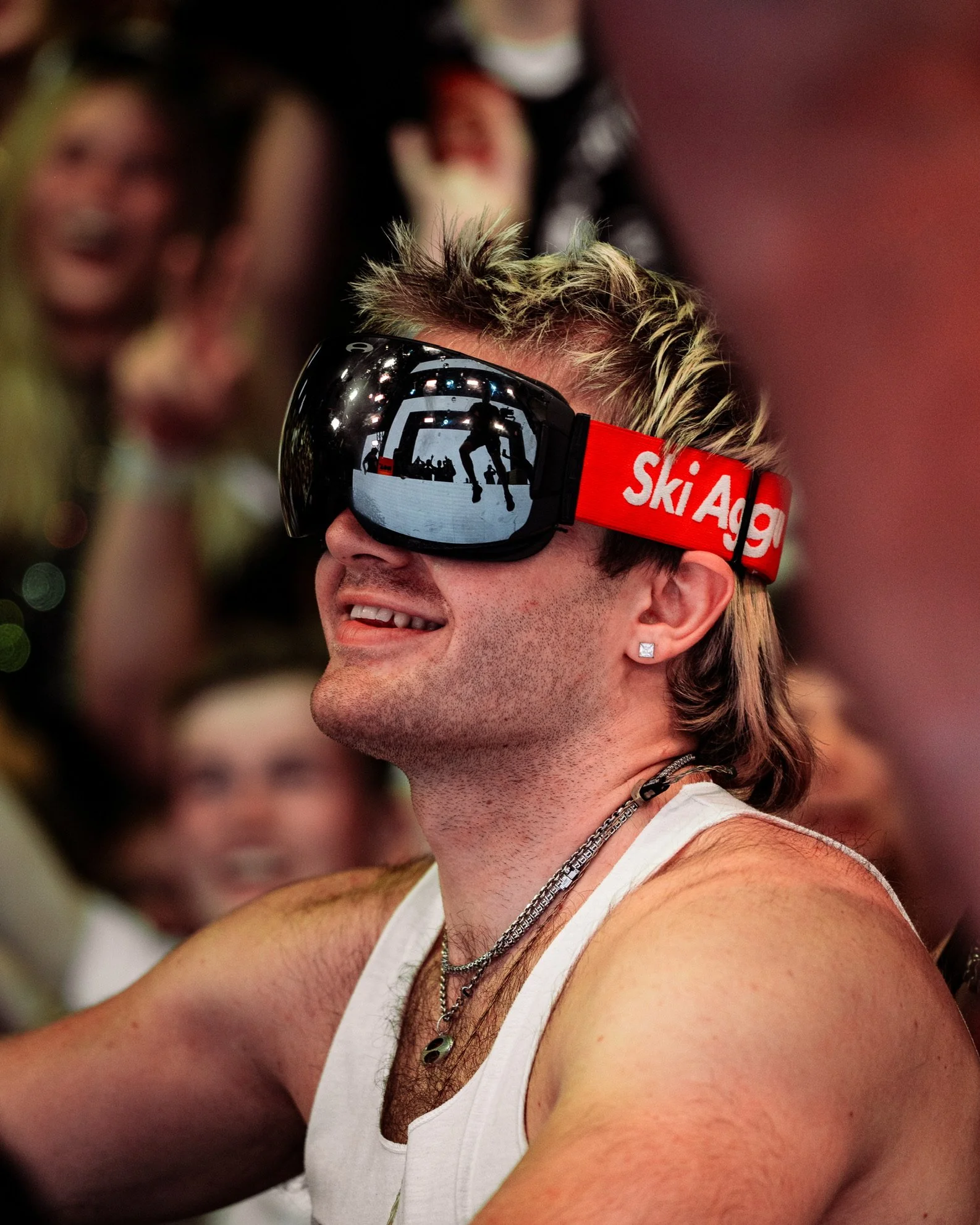 Ein lachender Mann mit durchgestyltem Haar trägt Skibrille mit reflektierendem Visier und zeigt Tattoos und Schmuck, im Hintergrund Menschen in einer Party-Umgebung.