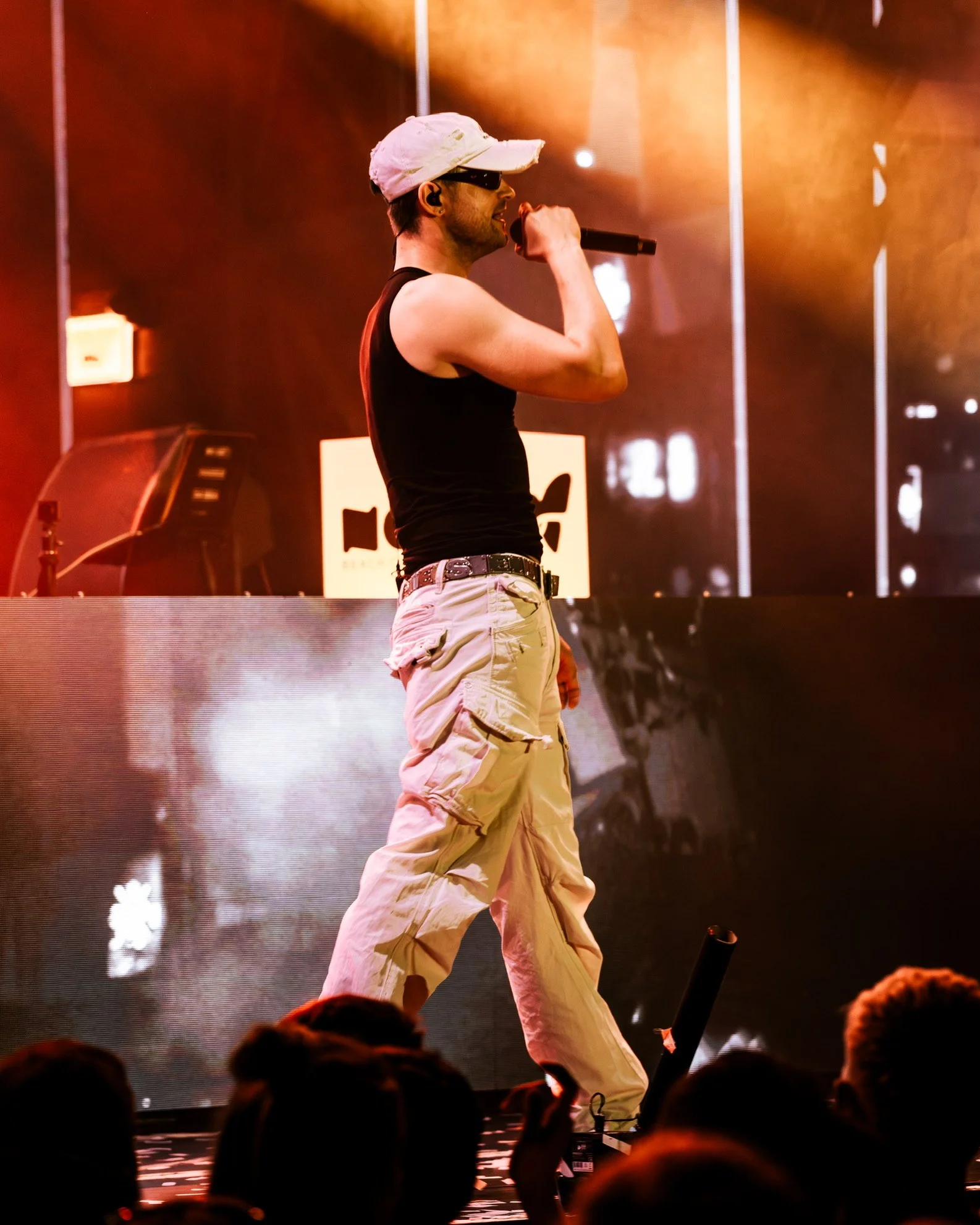 Ein männlicher Sänger trägt eine pinke Kappe, eine schwarze Weste, hellbeige Cargo-Hosen und surft auf der Bühne mit einem Mikrofon in der Hand, während im Hintergrund bunte Scheinwerfer leuchten.