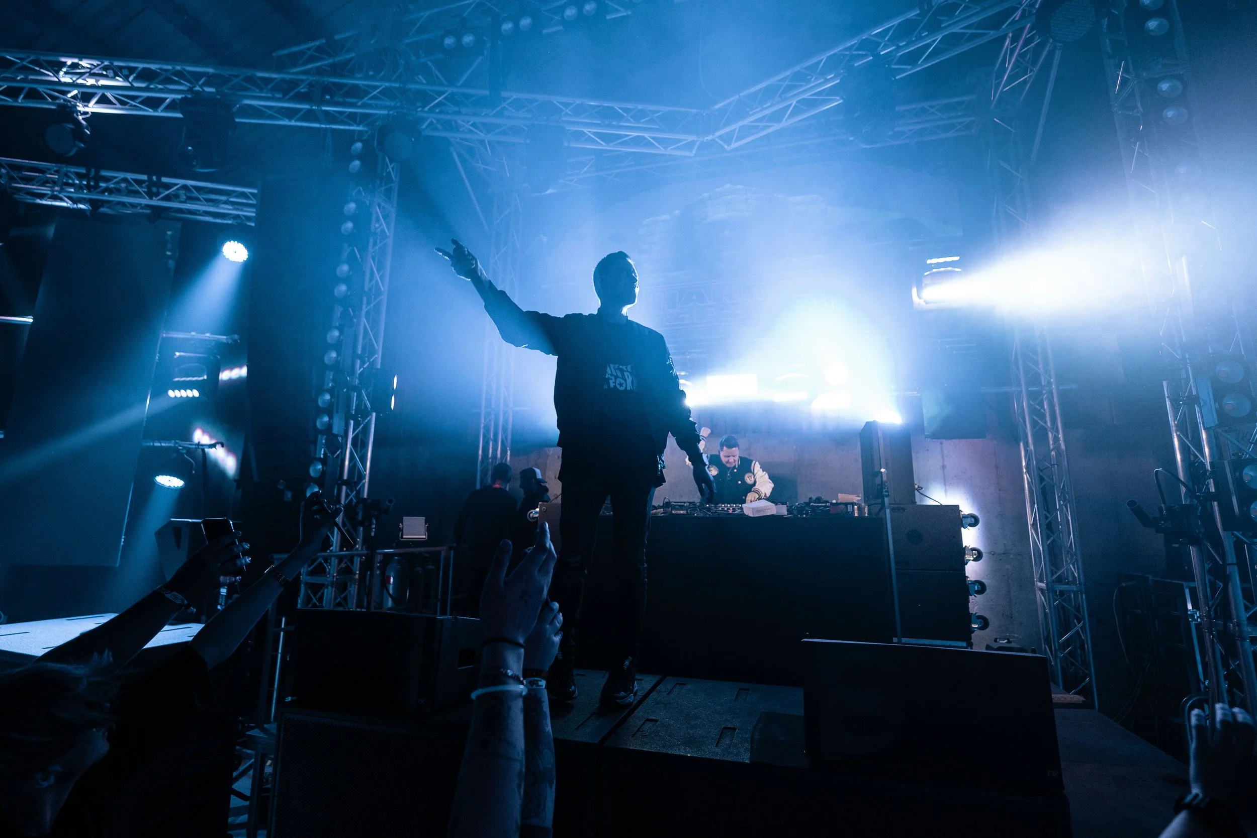 Ein DJ auf der Bühne eines Clubs oder Festivals, während er Musik macht, mit blauen Lichtern und einem Publikum, das Fotos macht.