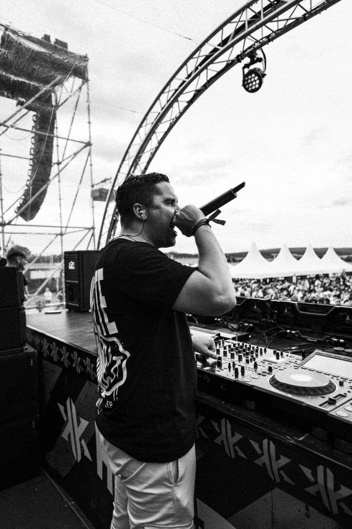 Ein DJ performing auf einer Bühne, während er in ein Mikrofon singt, im Hintergrund ein Publikum bei einem Musikfestival unter Zelten, im Freien bei bewölktem Himmel. Tänzer und Lautsprecher sind sichtbar.
