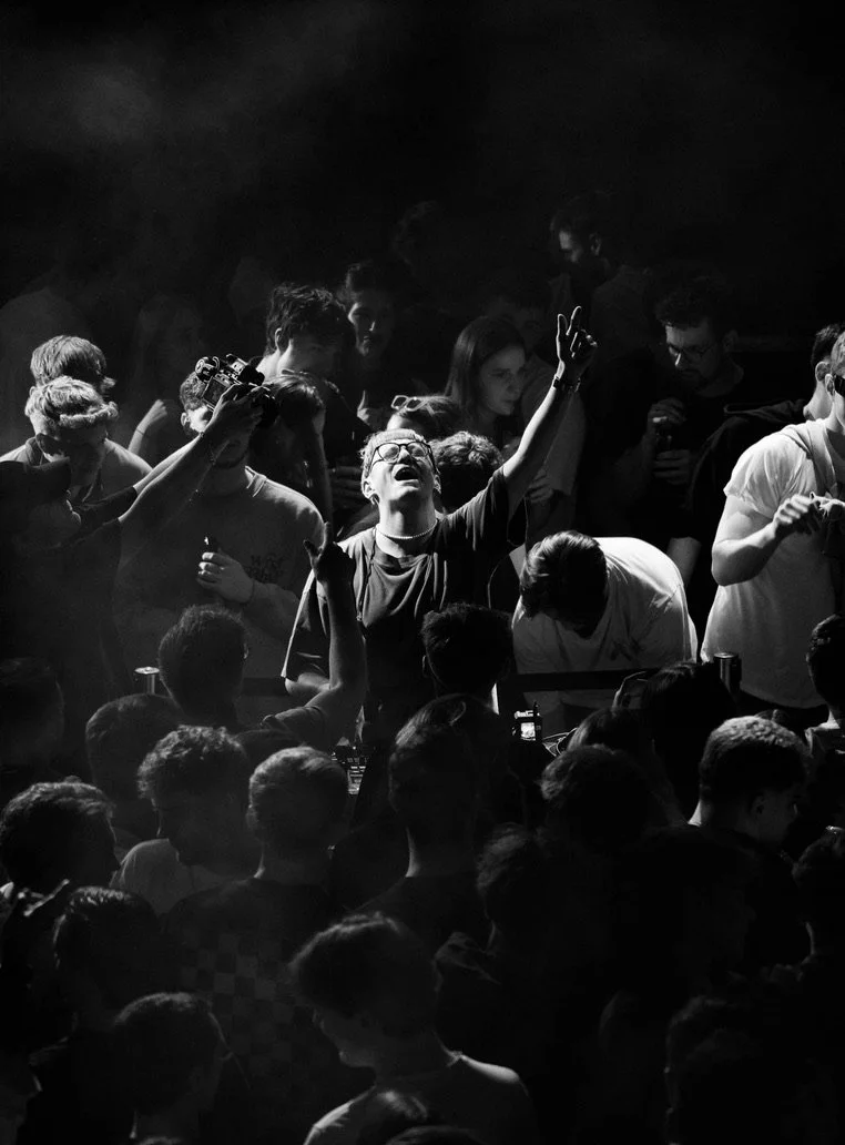 Eine Gruppe von Jugendlichen in einem dunklen Raum, einige scheinen zu feiern oder sich zu begeistern, mit einer Person in der Mitte, die hoch erhoben die Hand hebt.