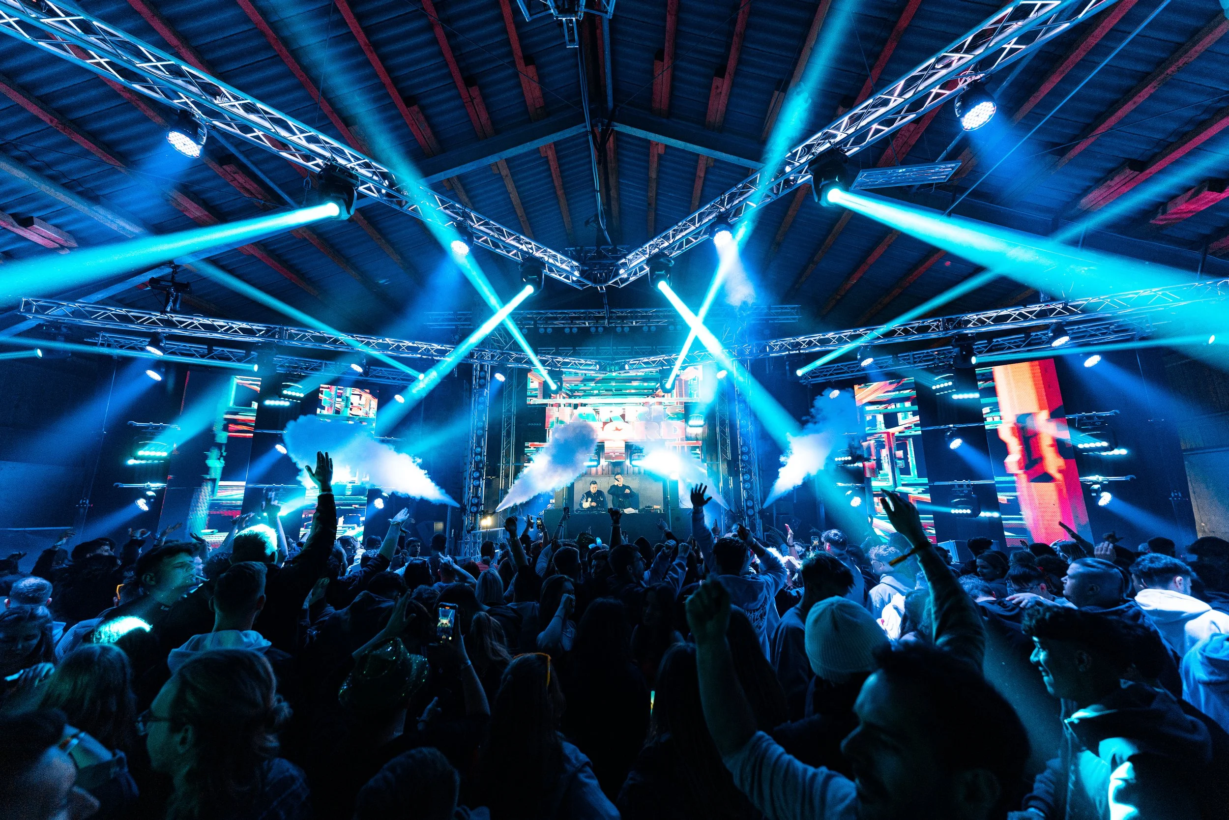 Menschen feiern bei einer elektronischen Tanzparty mit buntem Licht- und Lasershow in einer großen Halle.