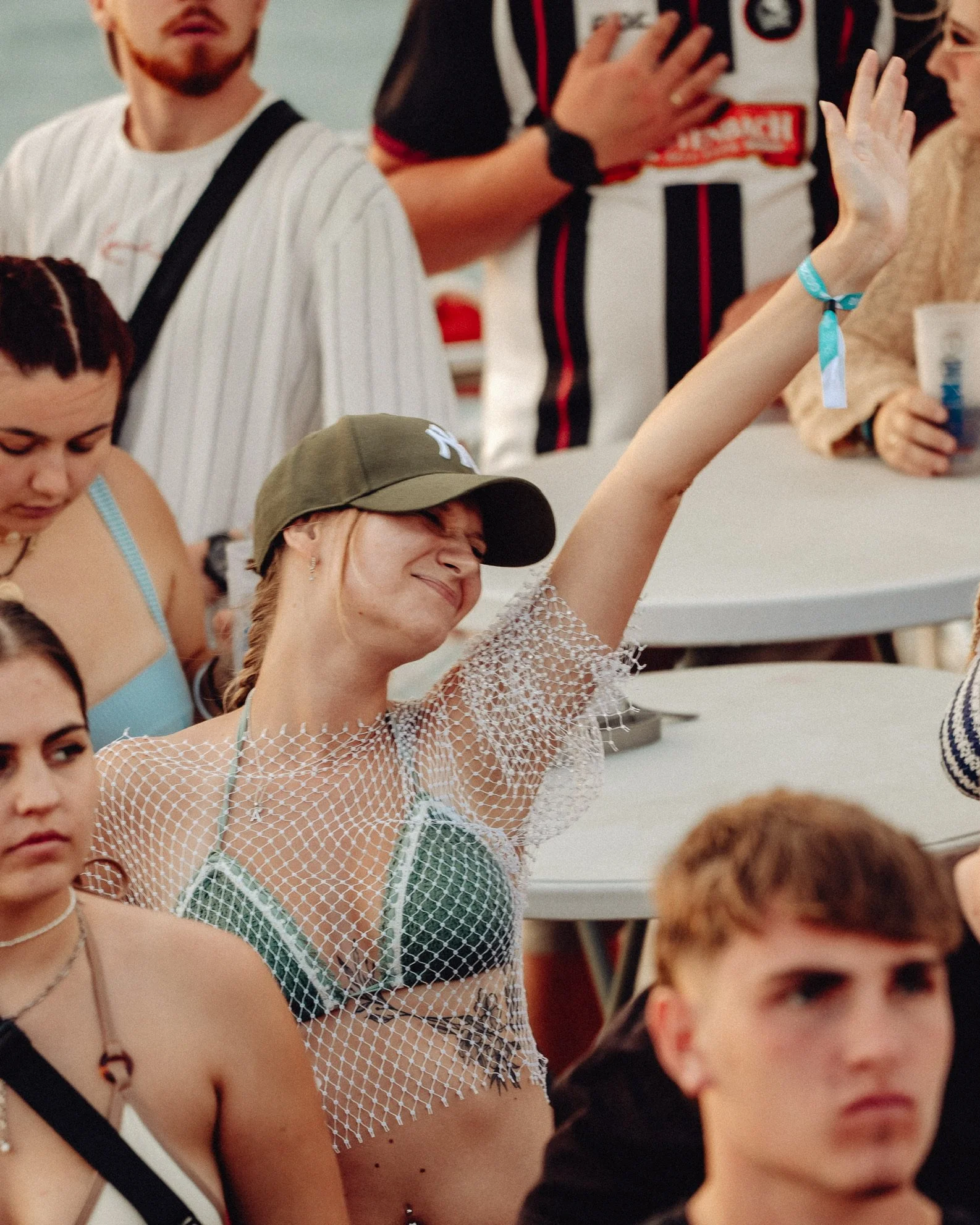 Eine junge Frau mit einem grünen Cap, einem Netzoberteil über einem Bikini, hebt die Hand und scheint zu tanzen oder zu feiern, während sie umgeben von anderen Jugendlichen ist.
