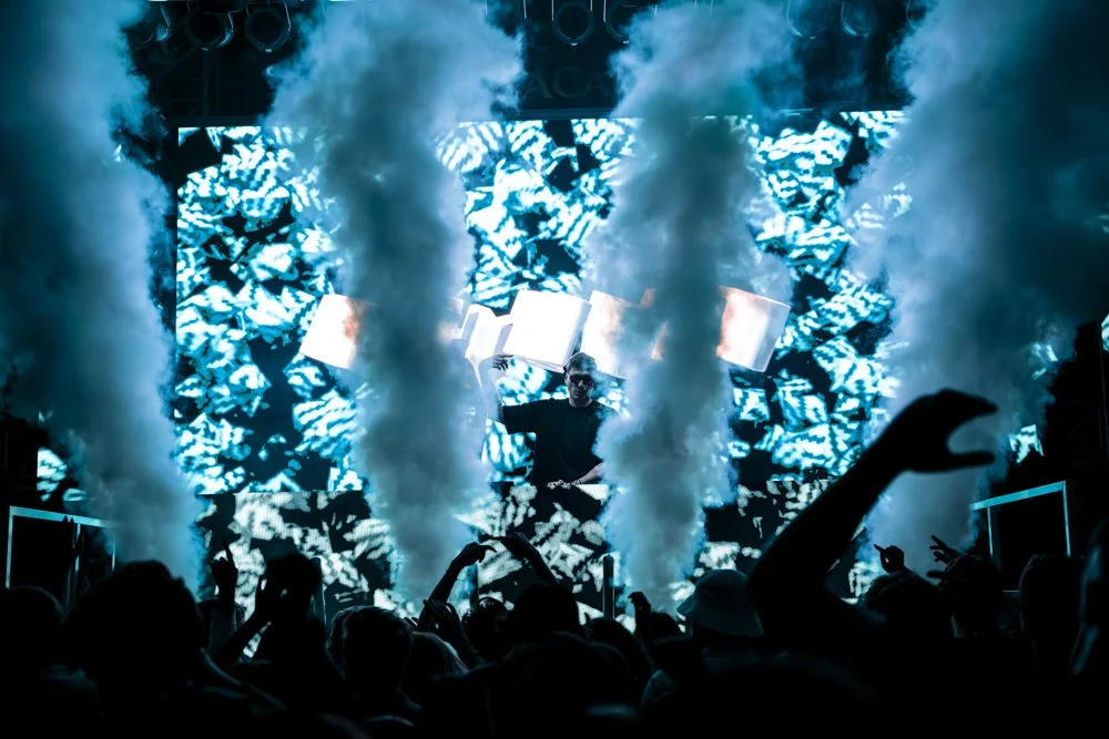 DJ auf einer Bühne bei einem Konzert, umgeben von Rauch und einer großen, leuchtenden LED-Wand mit abstrakten Mustern, Publikum im Vordergrund.