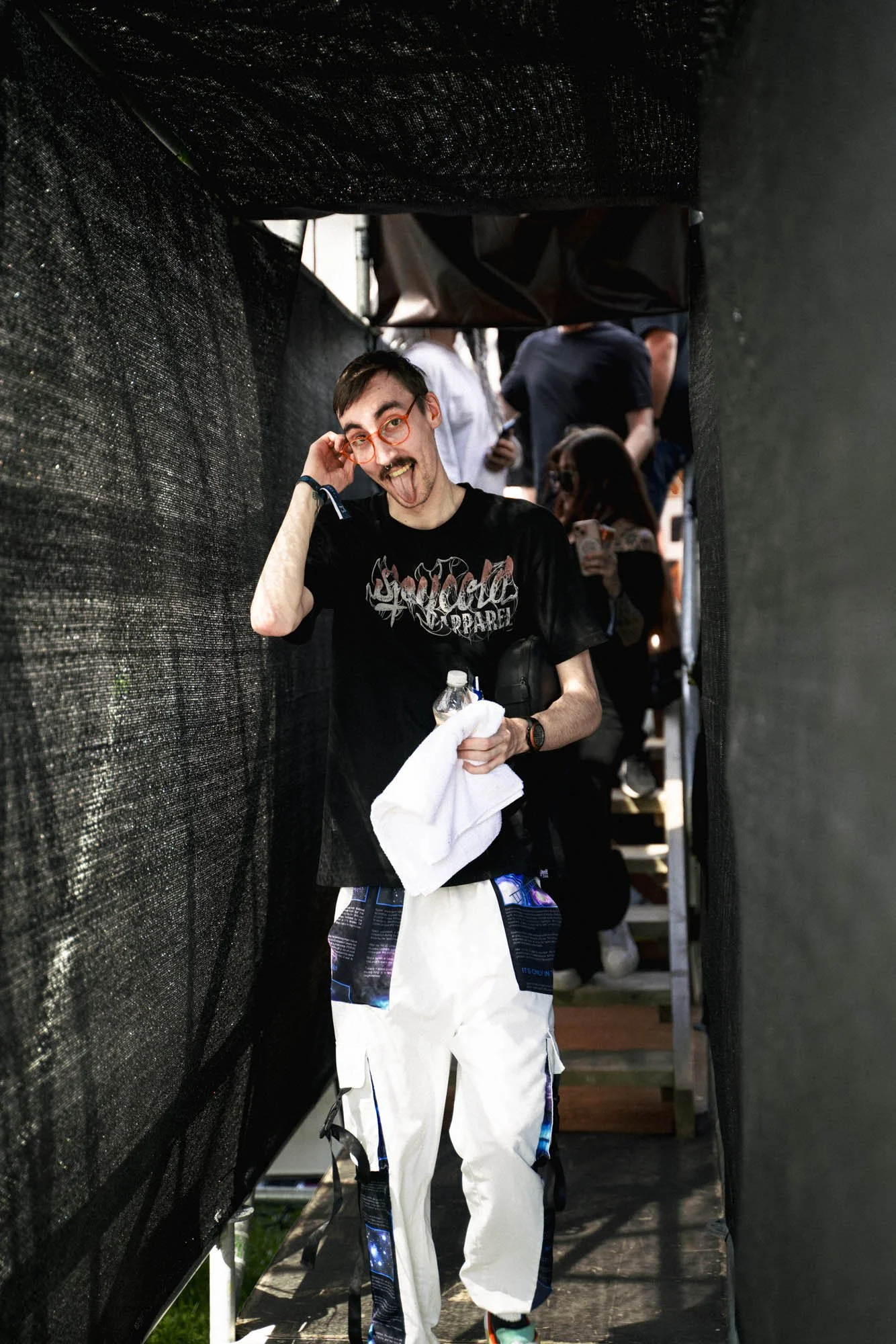 Junger Mann, der einen roten Brillenträger trägt, macht ein lustiges Gesicht und streckt die Zunge heraus, während er durch eine dunkle Passage geht, umgeben von anderen Menschen.