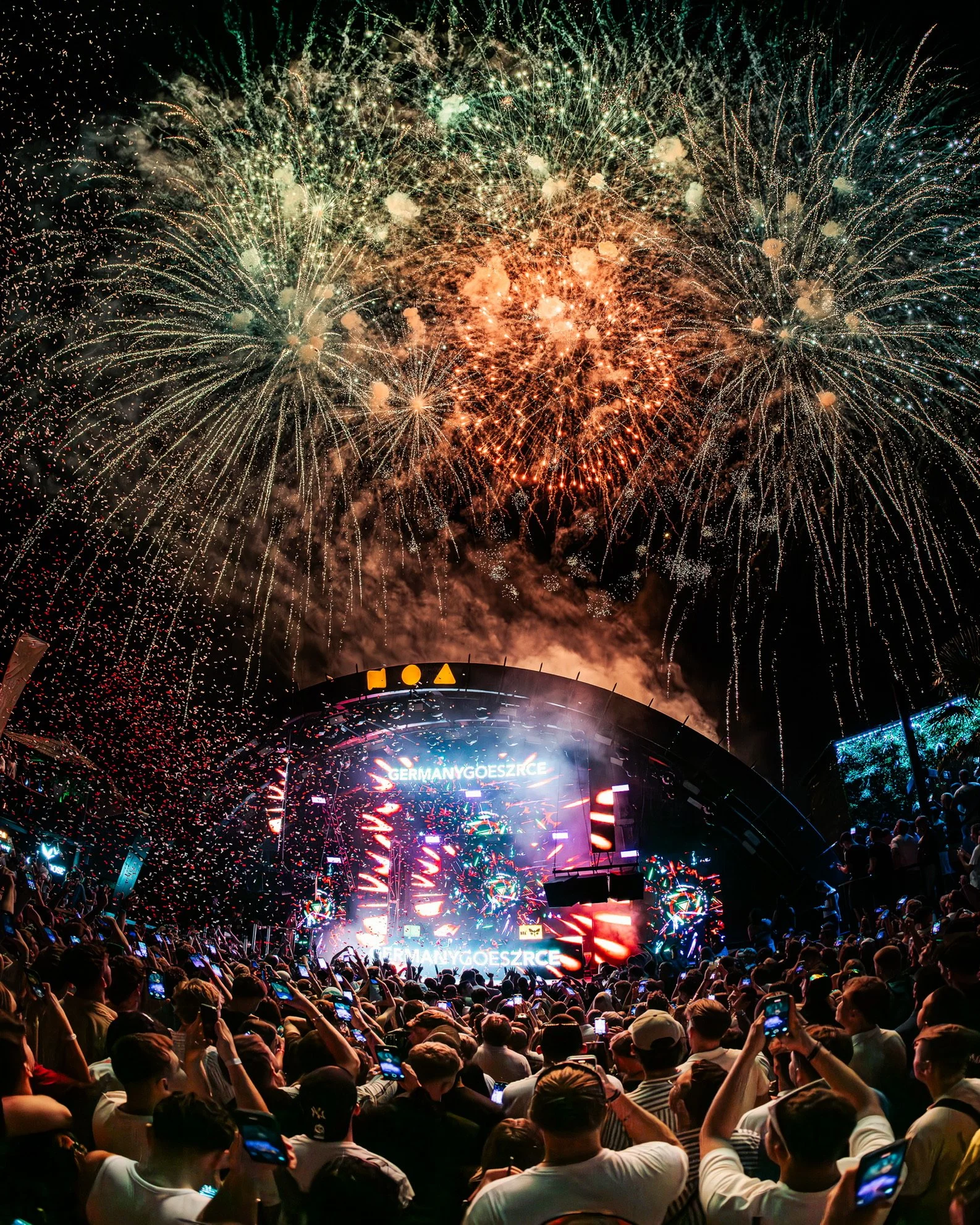 Feuerwerk über einer großen Menschenmenge bei einem Konzert oder Festival, mit einem Bereich in der Mitte, auf dem 'GERMANY GOES ZRCE' auf LED-Bildschirmen steht.