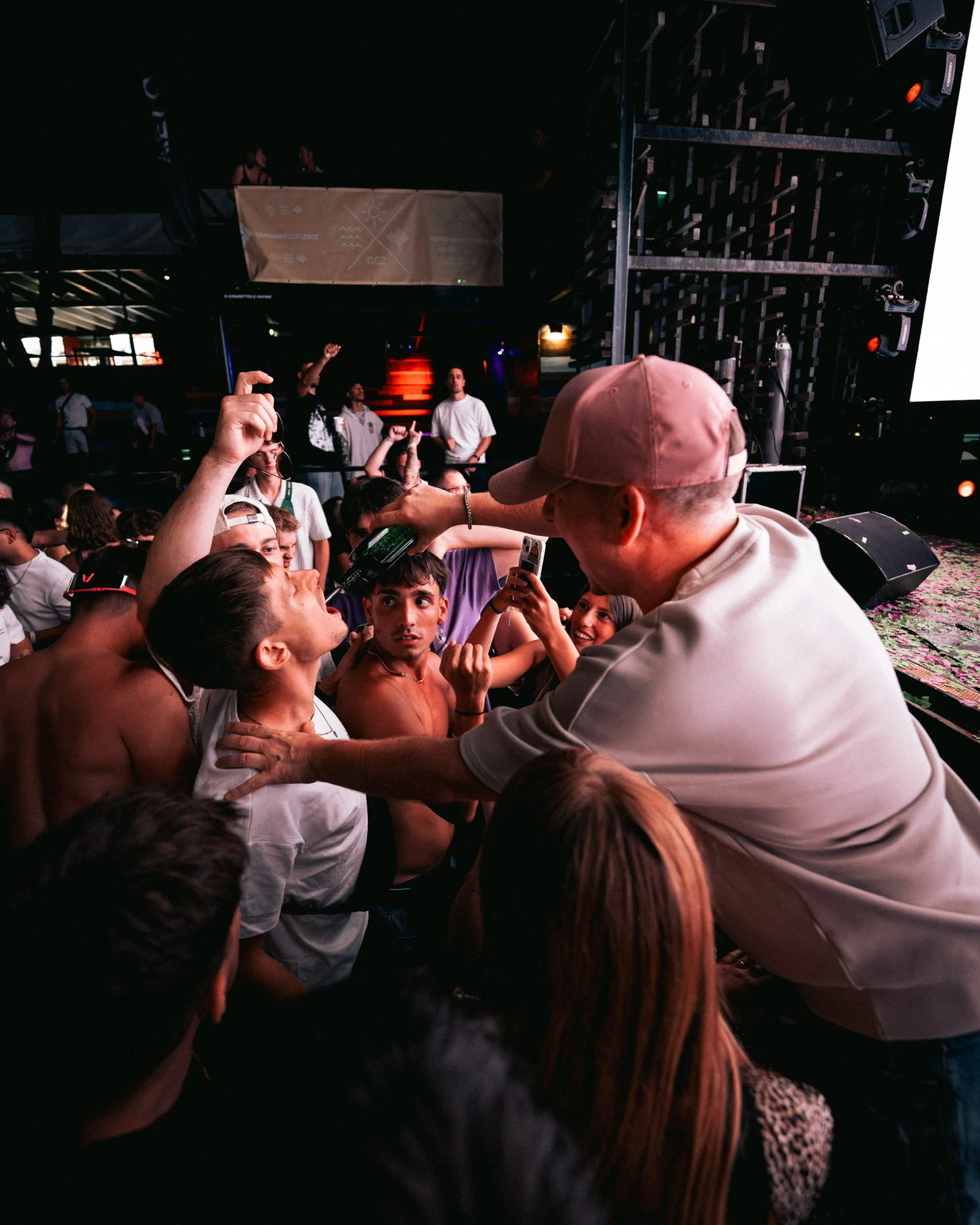 Mensch beim Feiern auf einer Party, hält eine Flasche und verteilt Getränke an die Gäste, bei schummrigem Licht in einem Club oder Konzertsaal.