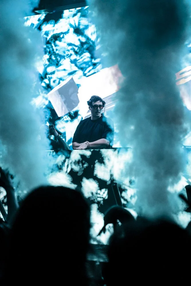 Ein DJ in dunkler Kleidung mit Sonnenbrille hinter einem Mischpult, umgeben von blauen Rauch- und Lichteffekten bei einem Konzert oder Clubveranstaltung.