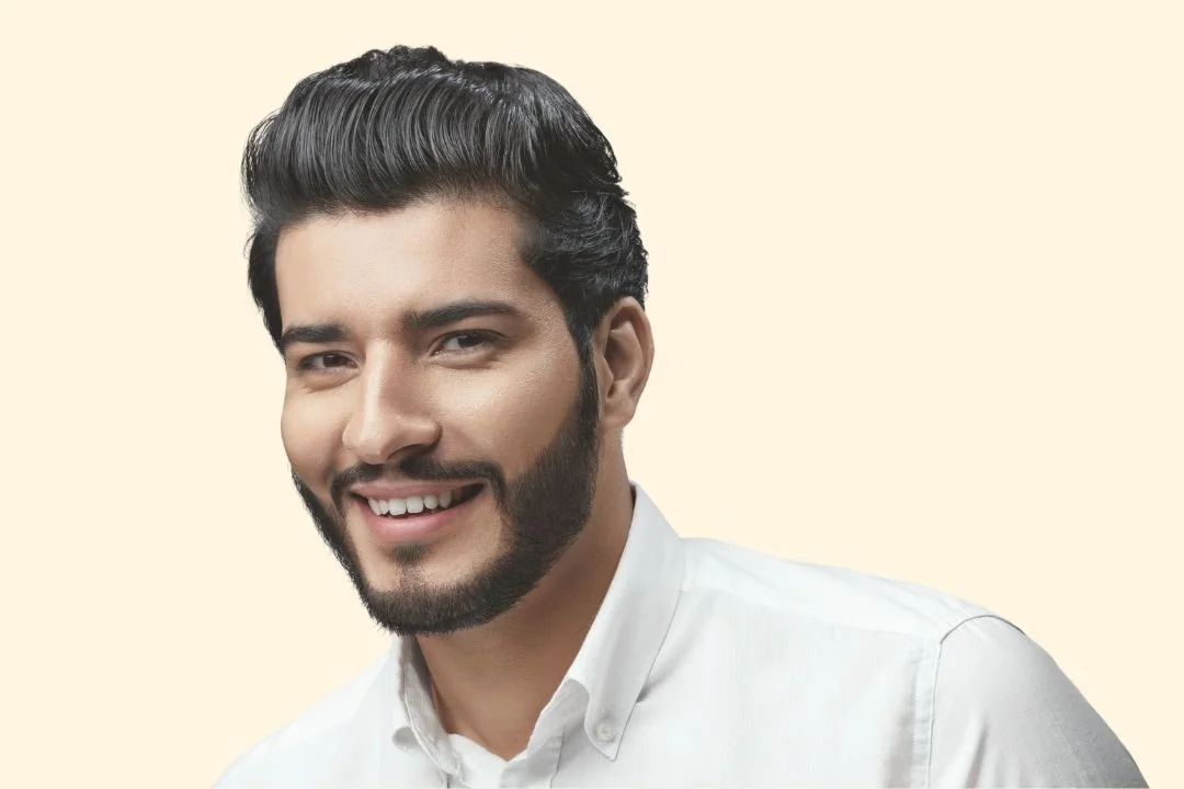Headshot of a smiling young man with dark, wavy hair and a beard, wearing a white shirt against a light beige background.