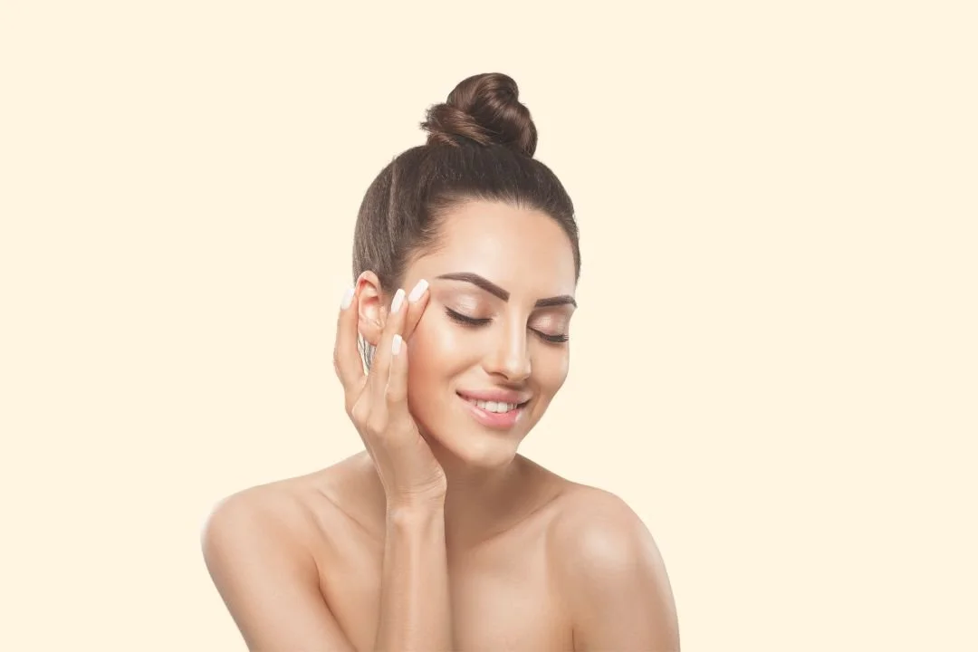 A woman with her eyes closed, smiling gently, touching her face, with her hair in a top knot.