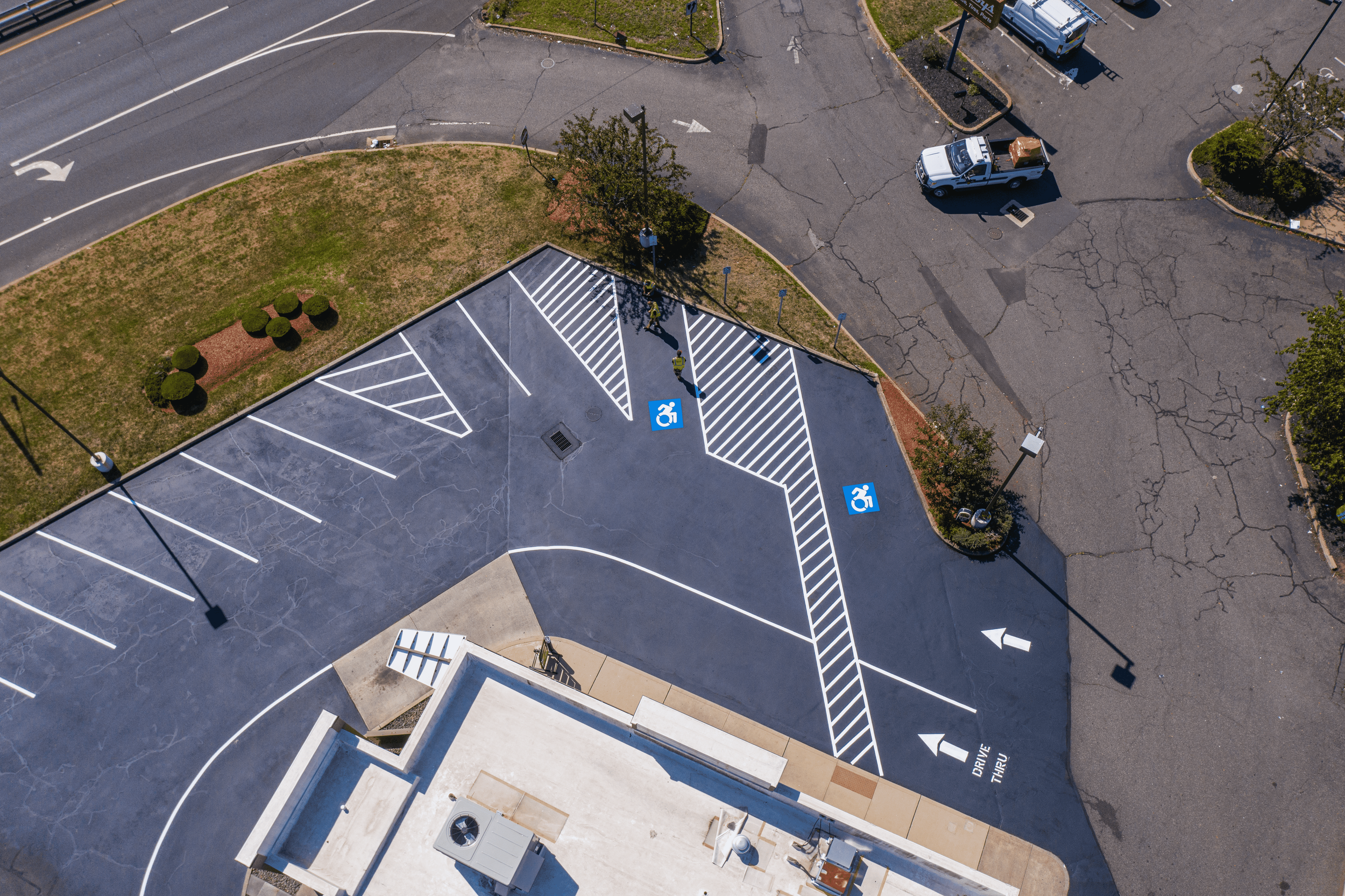 A parking lot with designated spaces including two blue handicap spots. The lot is adjacent to a grassy area with bushes and trees. There are two workers in yellow vests near the handicap spots. The surrounding asphalt has visible cracks and there ar