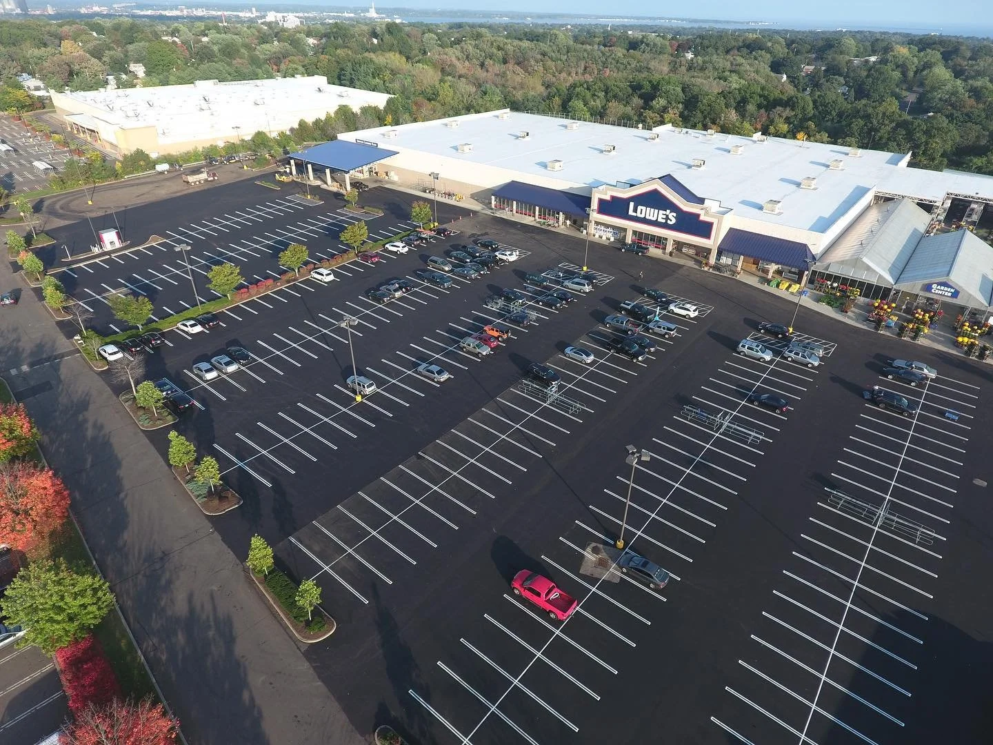 Empty parking lot outside Lowe's home improvement store with a few cars parked, surrounded by trees and a distant view of cityscape.