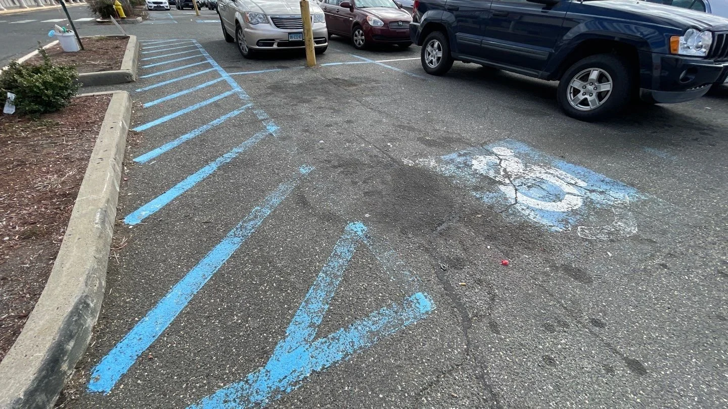 View of a parking lot with blue-striped accessible parking spaces, some of which are faded and cracked, adjacent to parked cars and a landscaped area.