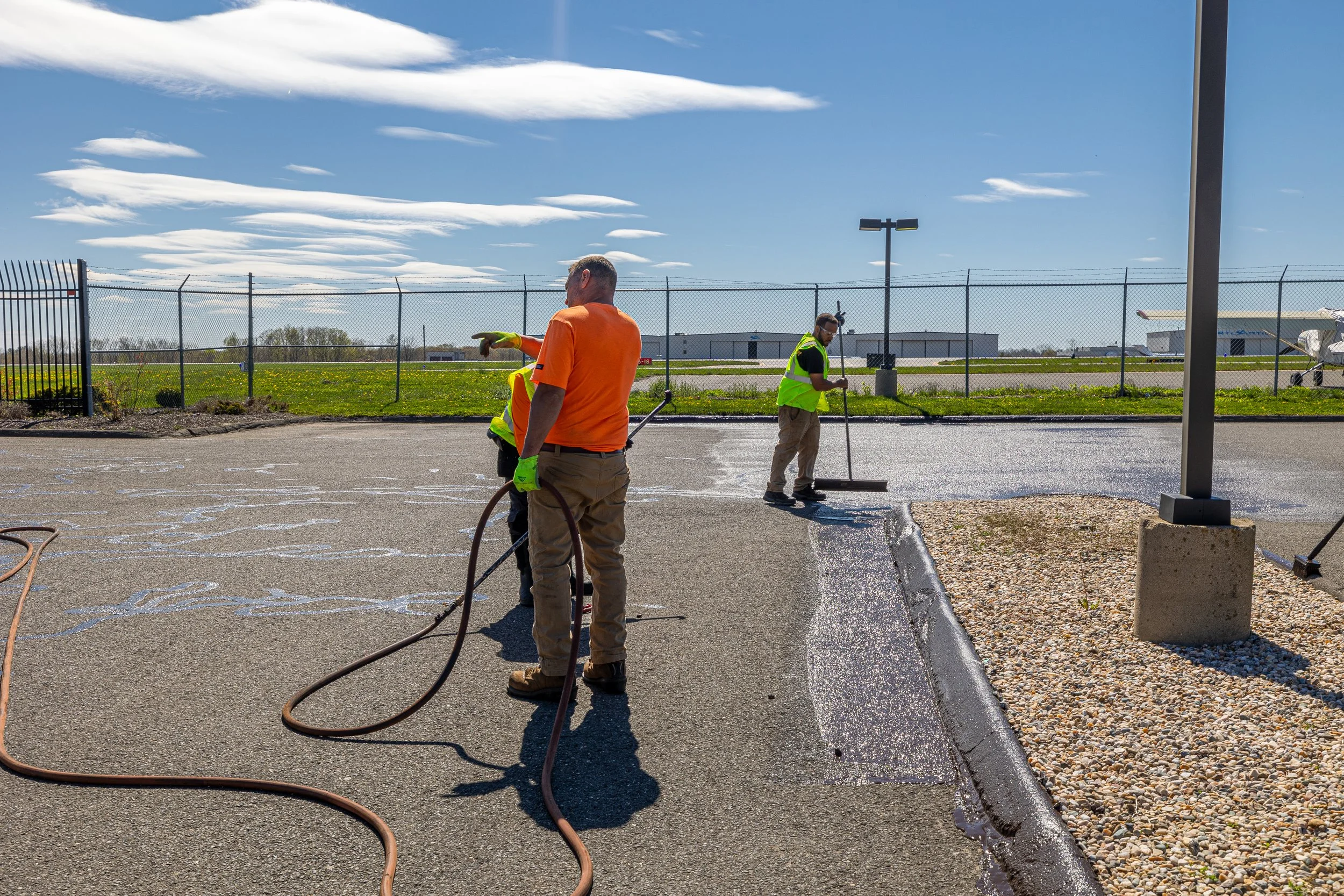 Workers sealing a parking lot with asphalt using a roller and hoses on a sunny day.