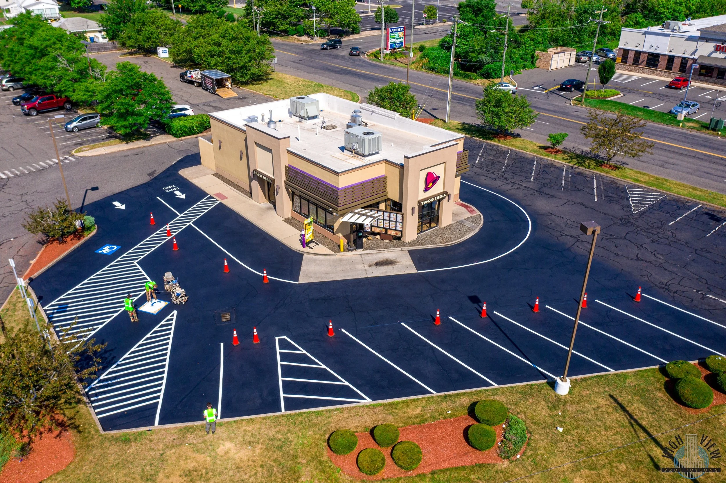 A newly paved parking lot with designated spots, some lined for handicap parking, and traffic cones set up. There are workers in safety vests painting or finishing markings near the entrance of a fast-food restaurant named Taco Bell. The building is 