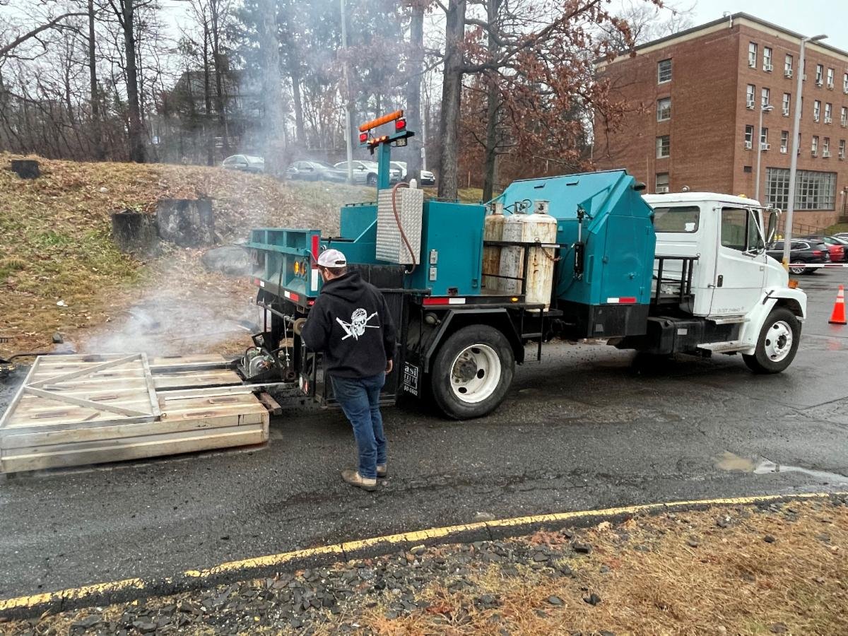 Person standing next to a street sweeper truck that is cleaning curbside debris as smoke or steam rises nearby.