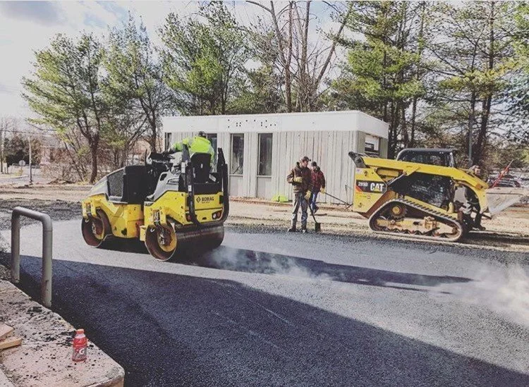 Construction workers paving the road with heavy machinery on a clear day, with trees and a small building in the background.