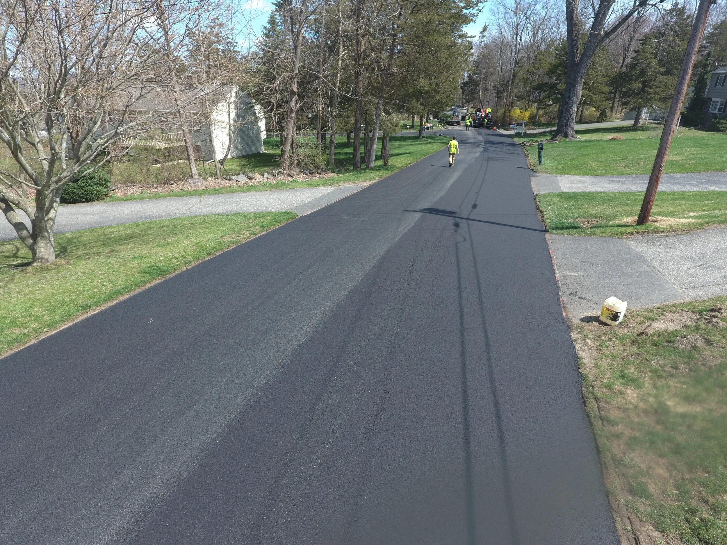 A residential street with new asphalt being laid. Visible workers and equipment at the distant end of the street, with a mailbox and trees along the sides.