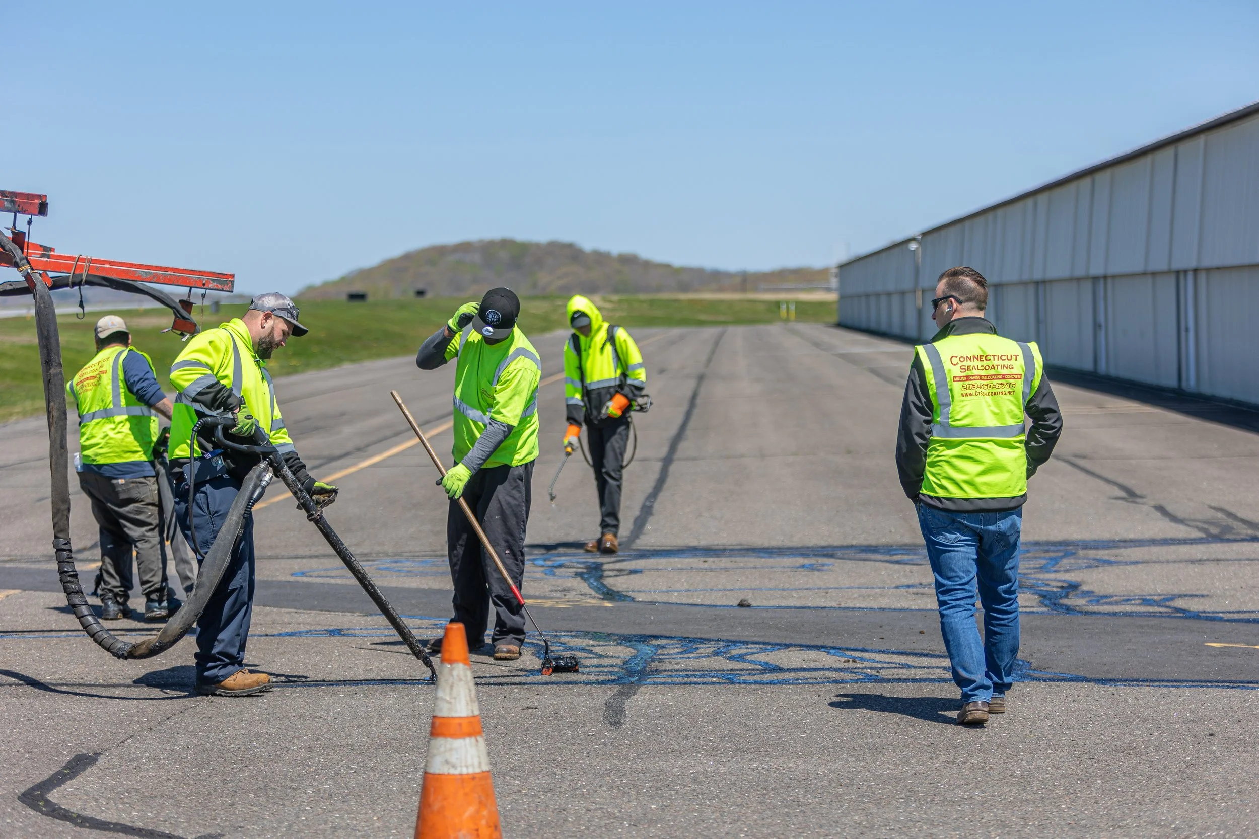 Workers in fluorescent safety vests repairing the asphalt on an airport runway with a strip of blue paint, while an orange traffic cone sits in the foreground and a large white building is in the background.