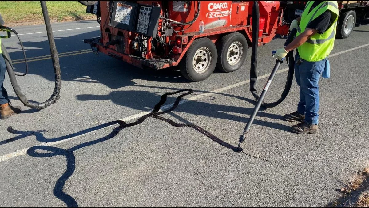 Workers repairing a crack in the road using specialized equipment with a red service vehicle in the background.