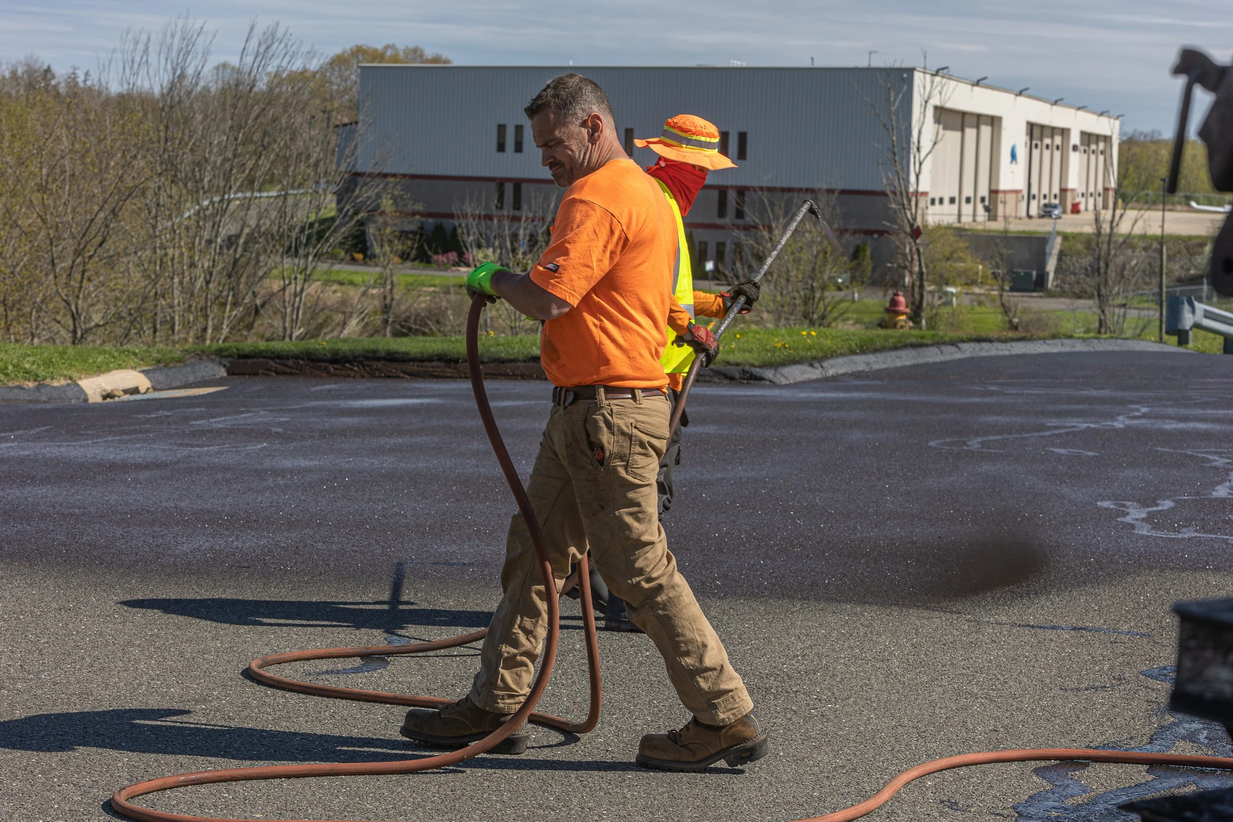 Two workers are applying a coating to an asphalt parking lot, one in an orange shirt and the other in a yellow vest, with the background featuring commercial buildings and trees.