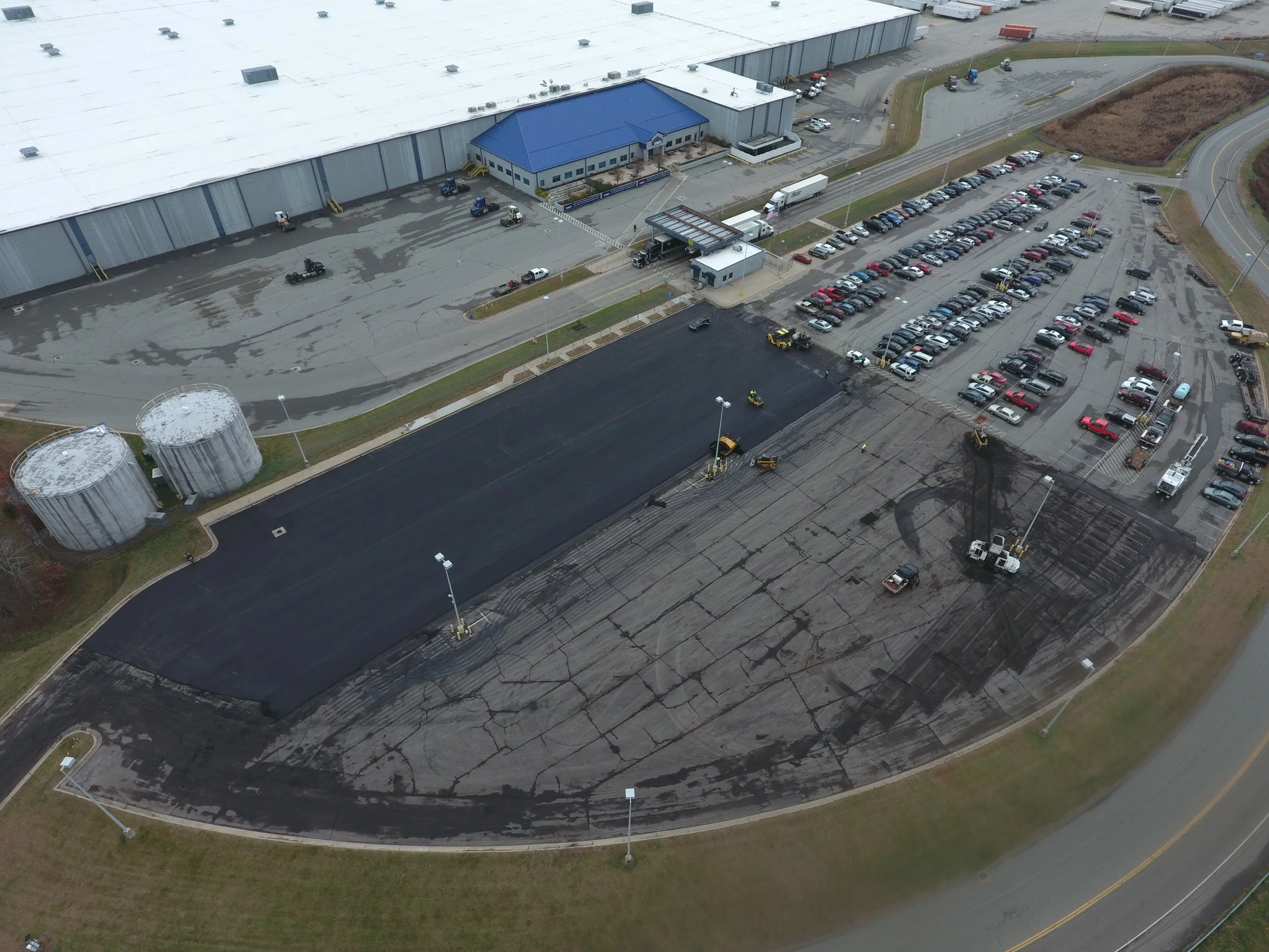 Aerial view of a parking lot under construction with sections of freshly paved asphalt, construction equipment, and parked vehicles, adjacent to a large industrial building.