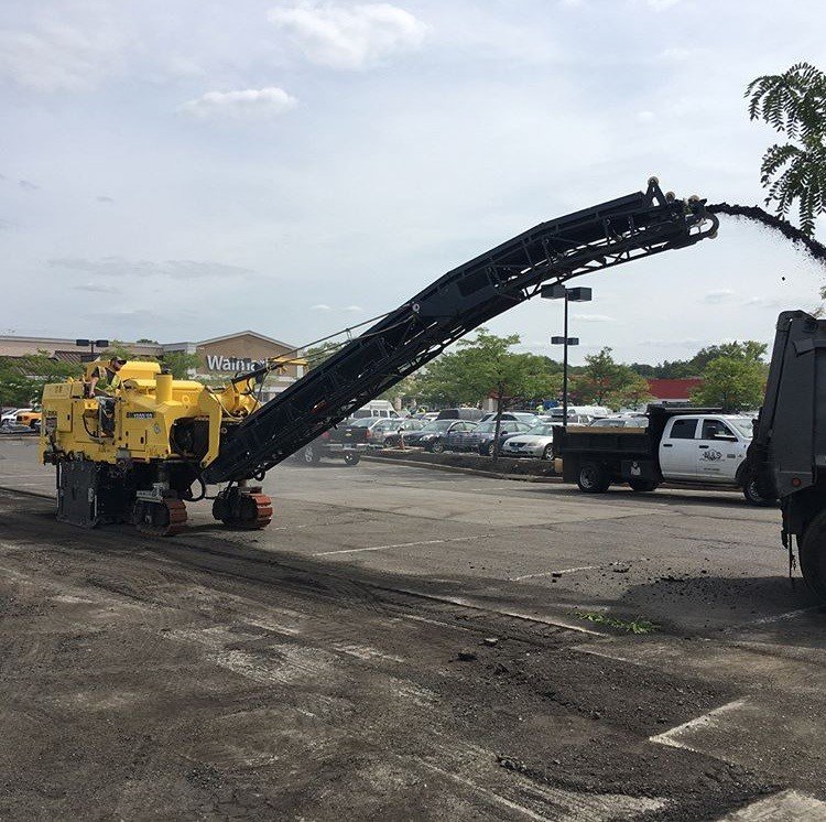 Street construction equipment working on pavement repair, with a parking lot and store in the background.