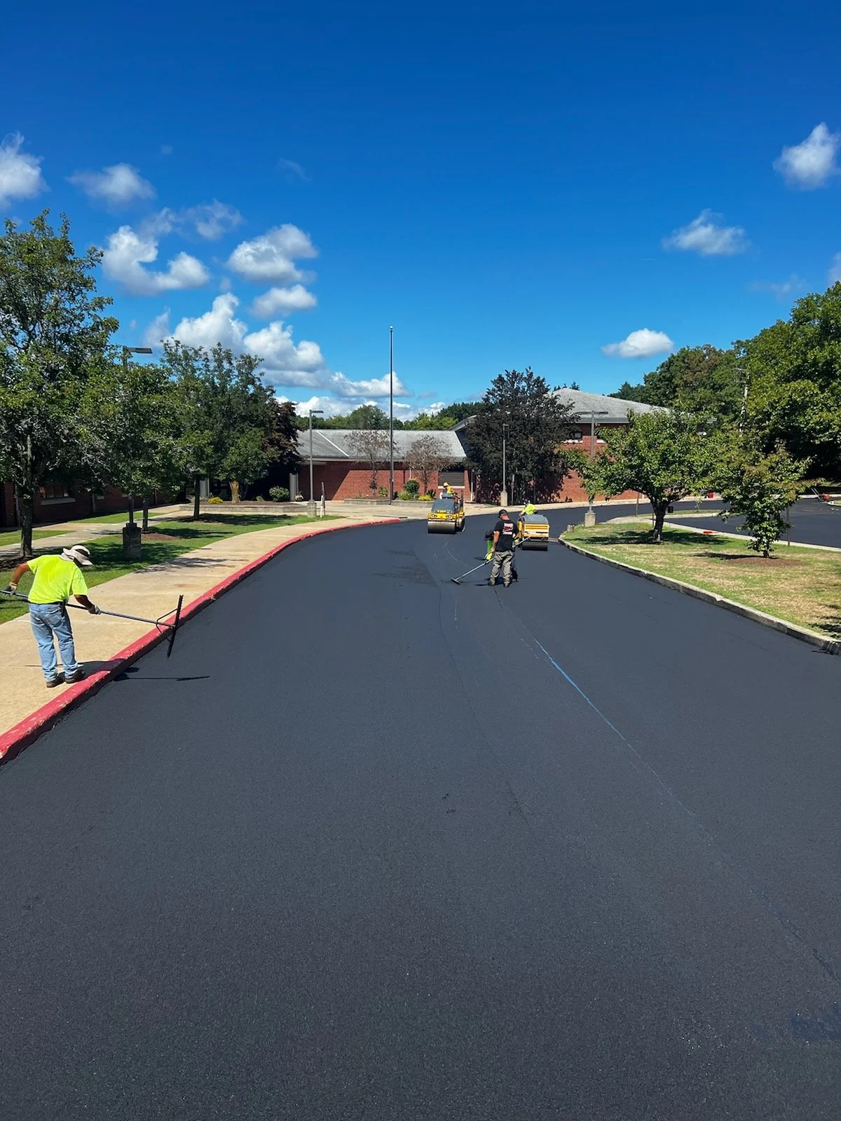 Workers paving asphalt on a curved road with trees and a brick building in the background.