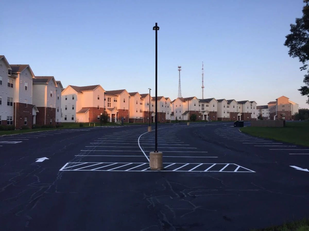 Empty parking lot in front of a residential apartment complex at dusk, with a lamppost in the center and few trees on the sides.