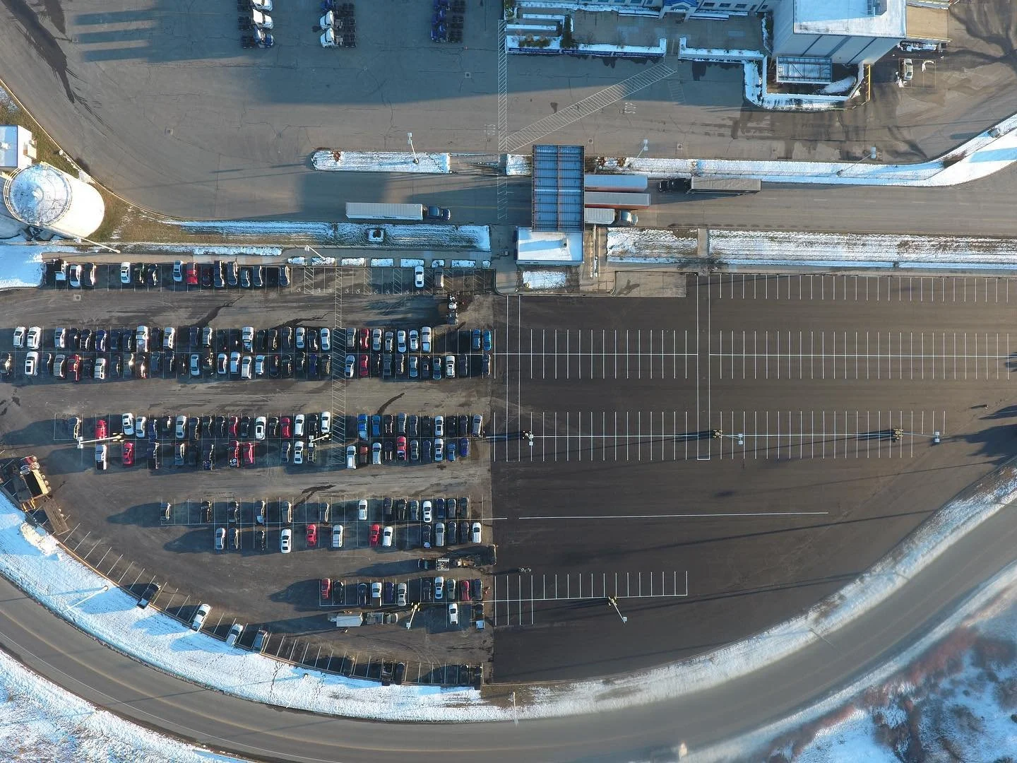 An aerial view of a parking lot with mostly empty spaces and a few cars parked along the edges. Snow is visible on the ground and surrounding areas.