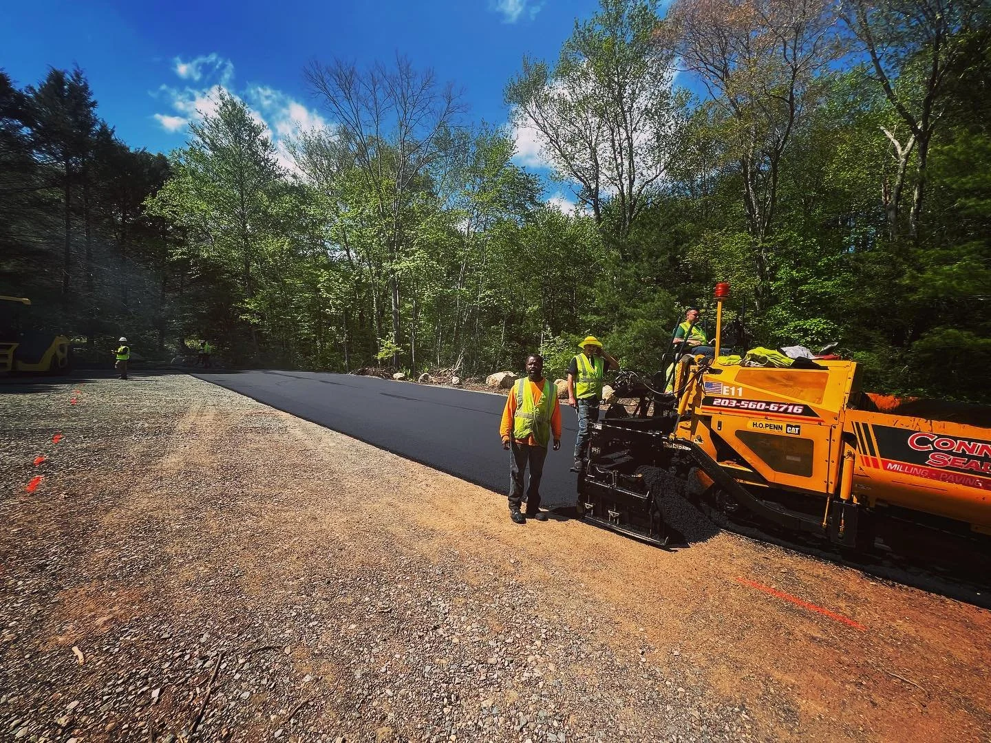 Workers laying new asphalt on a road in a wooded area, with trees and blue sky in the background.