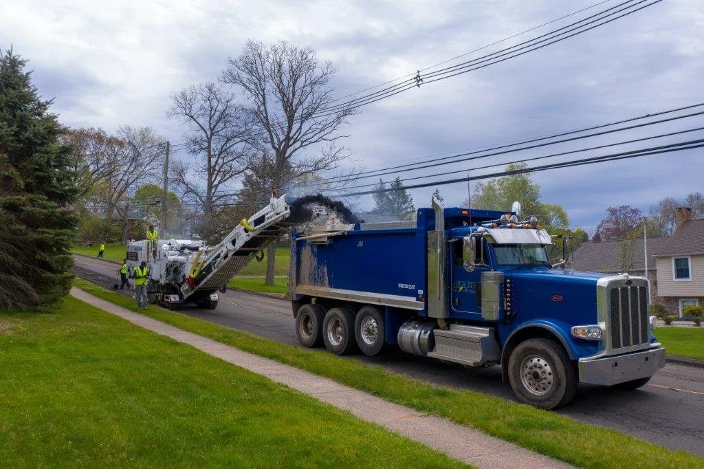  Several workers in safety vests are nearby, and there are trees and houses in the background.