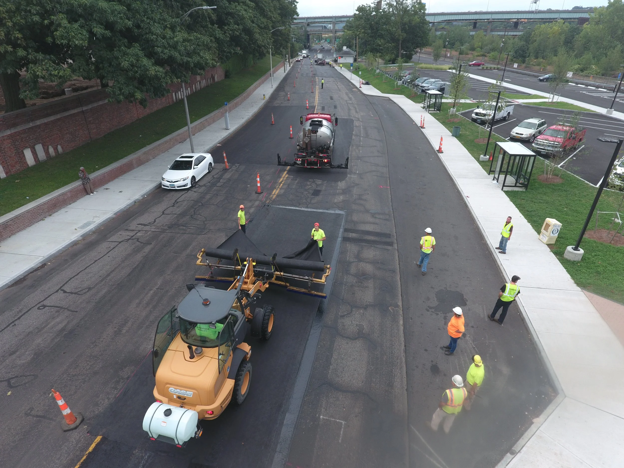 Construction workers and machinery resurfacing a street with asphalt, with traffic cones and parked cars visible.