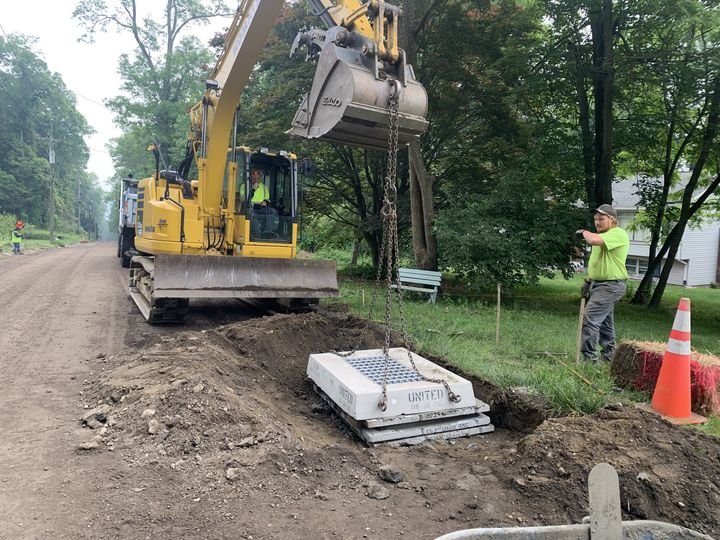 A construction site with a yellow excavator placing a large concrete block into a dug trench, with workers and orange traffic cones nearby.