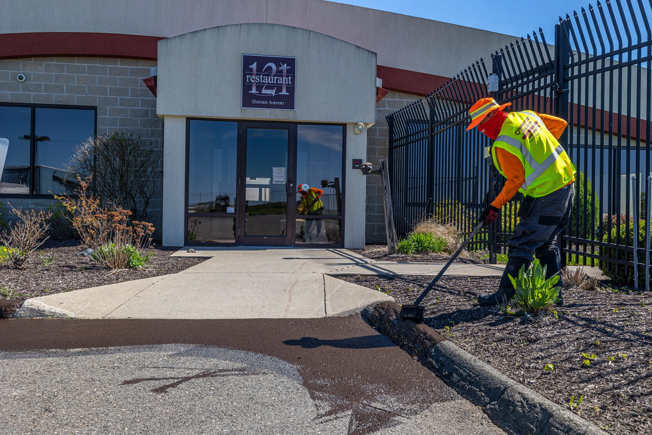 A worker in a high visibility vest and protective gear is laying new asphalt outside a building with a sign reading "10 restaurant Oxford Airport." The worker is using a tool to spread fresh asphalt along the edge of the sidewalk near a black metal f