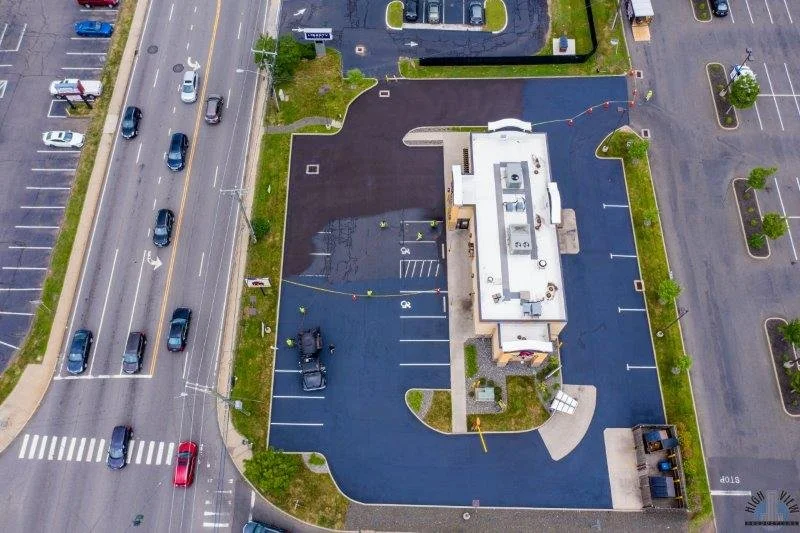 An aerial view of a commercial building surrounded by a parking lot with a few parked cars, adjacent to a busy multi-lane street with moving vehicles, along with some trees and green spaces.
