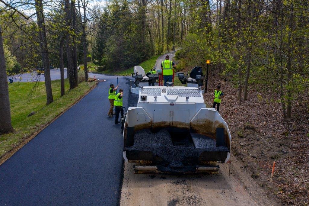 Workers guiding asphalt paving machine as it lays new asphalt on a rural road surrounded by trees.
